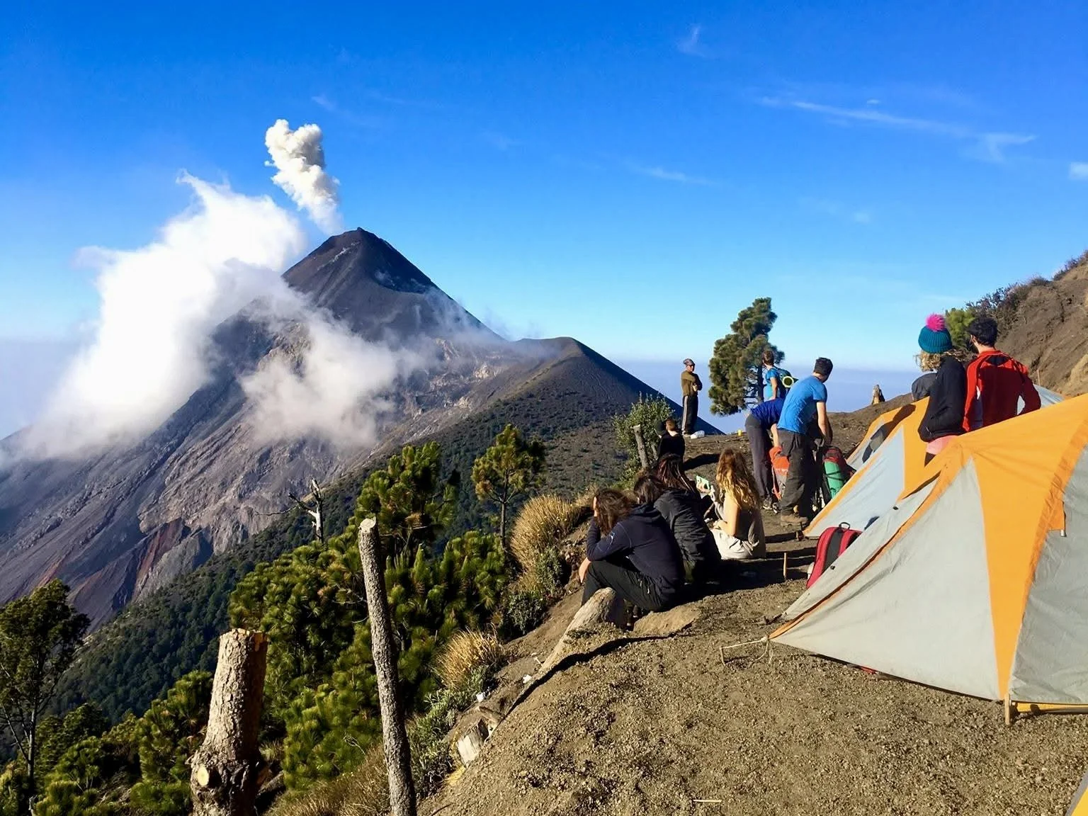 Multiple orange tents lined up on the edge of a mountain with people sitting on the ground looking at a neighboring erupting volcano during the day