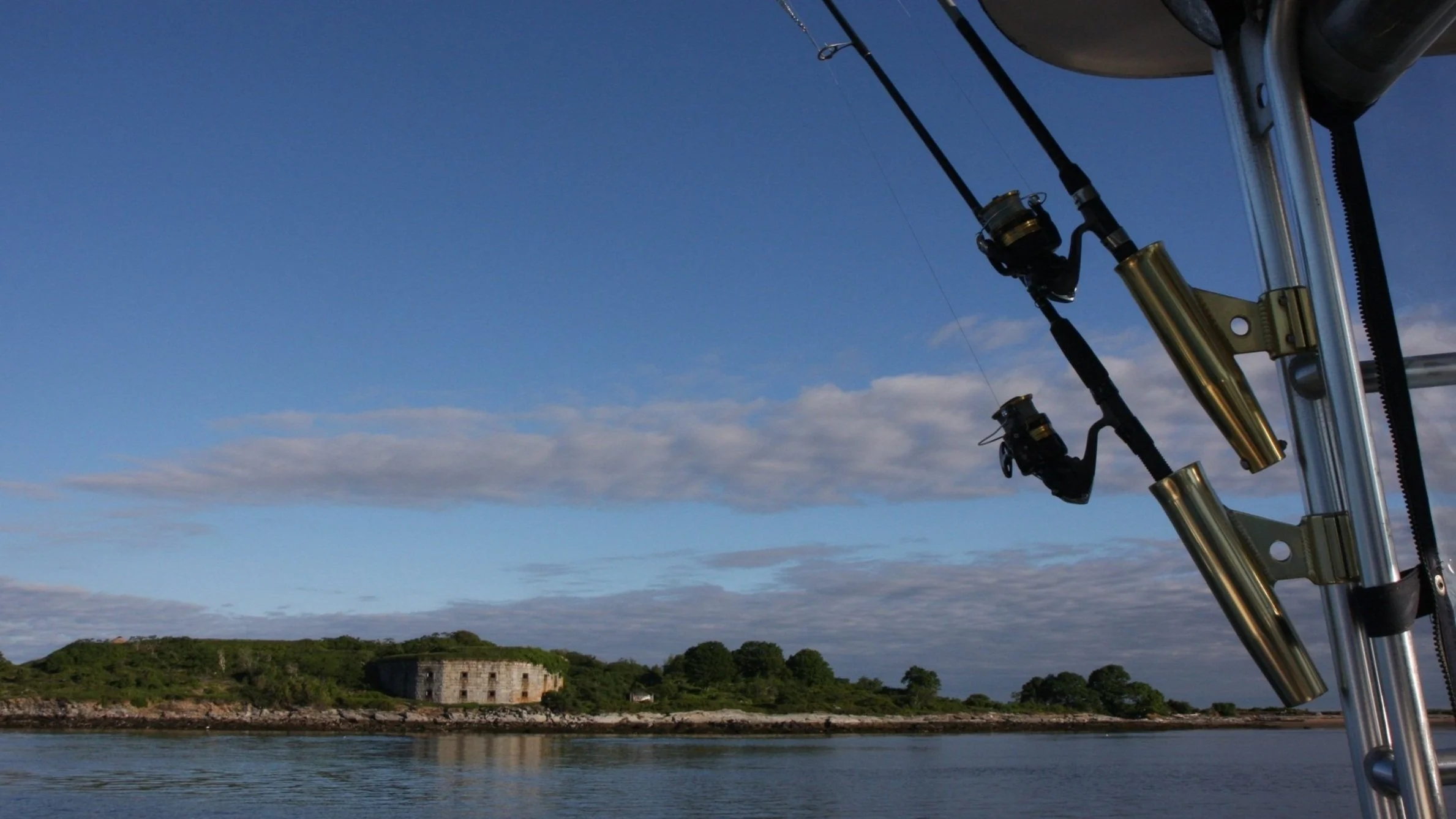 Fishing rods mounted on a boat with a view of a river, greenery, and an old building in the distance.