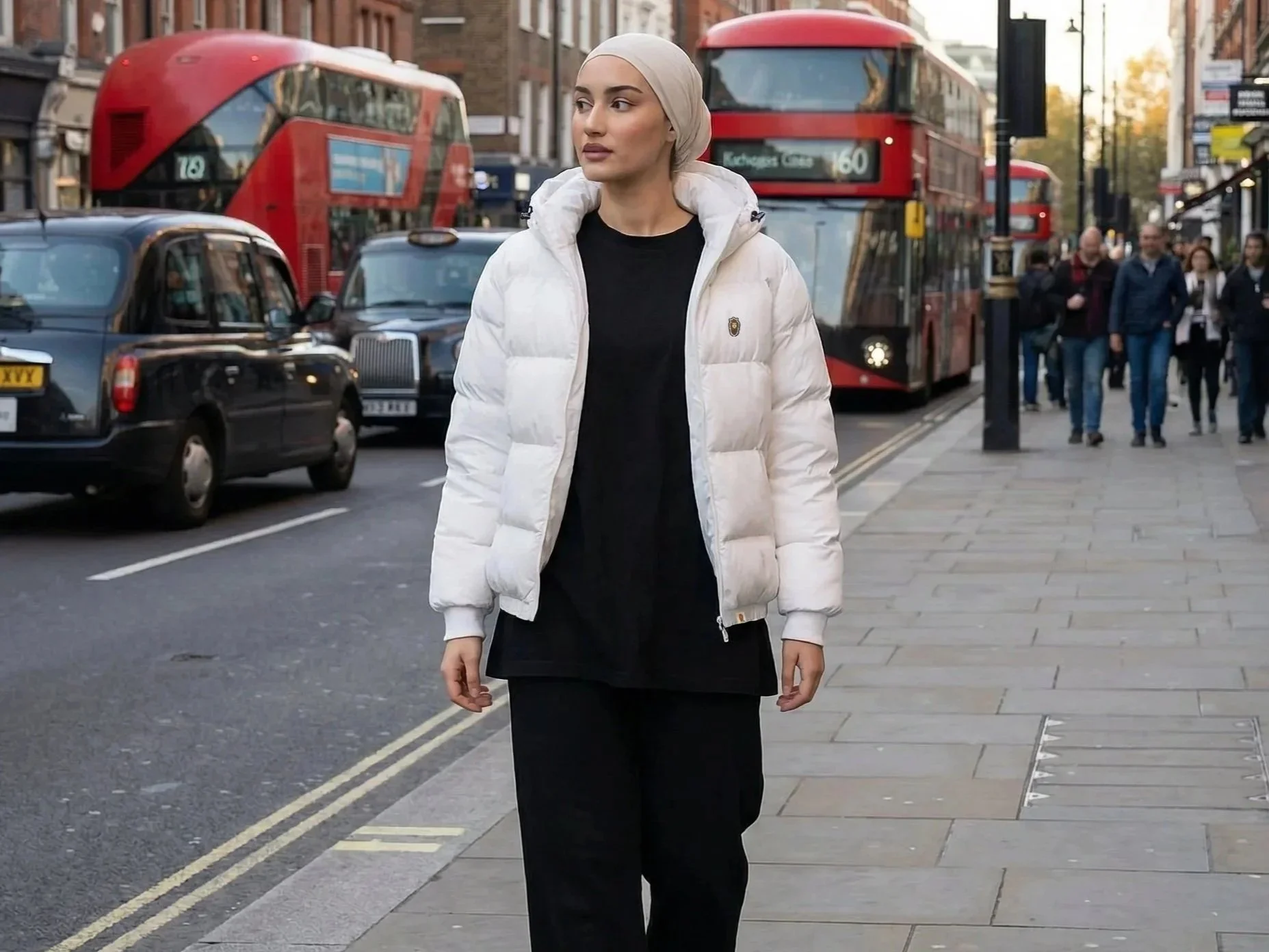 A woman in a beige hijab and white puffer jacket walking on a city sidewalk, with a red double-decker bus and cars in the background.
