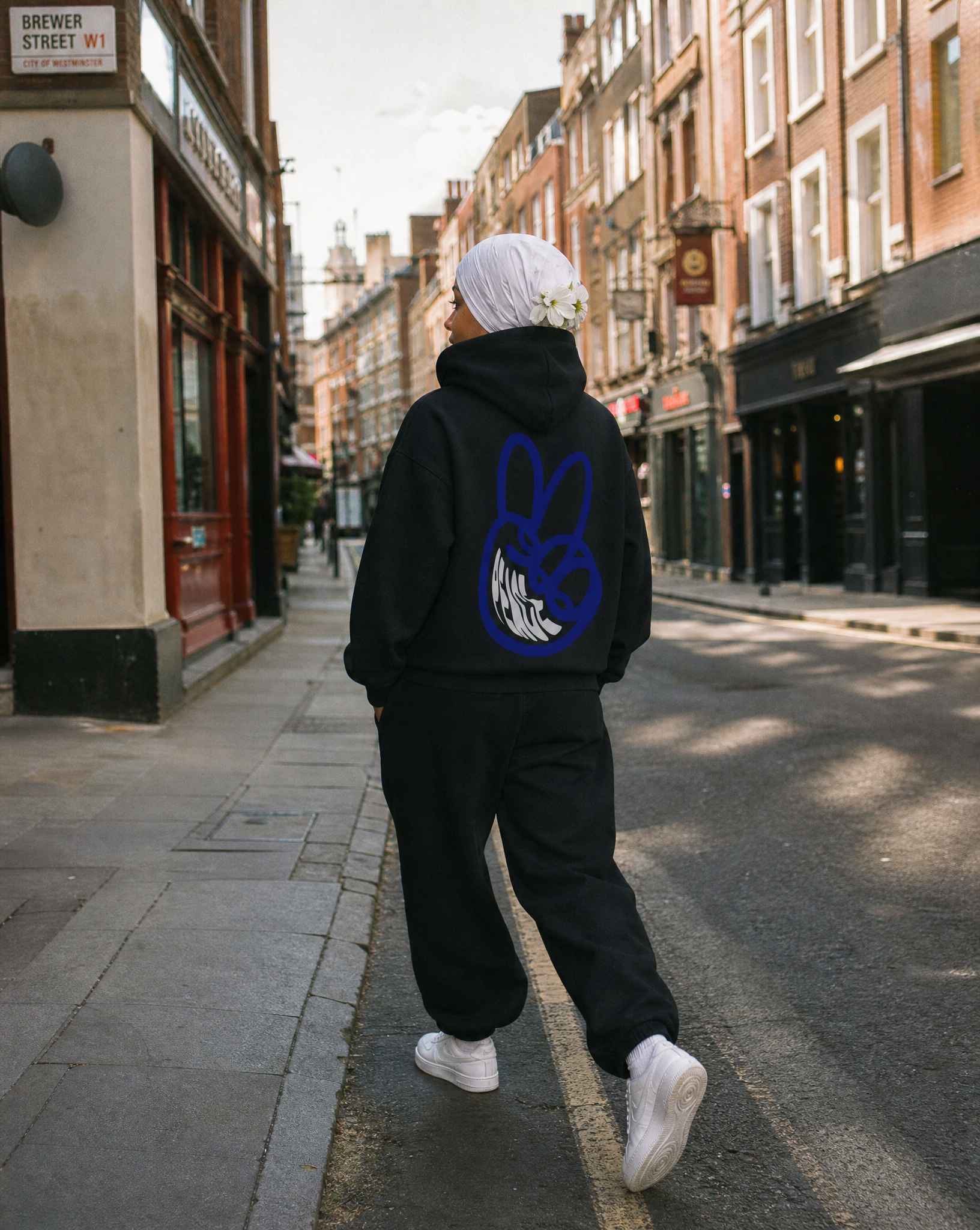 Person walking on a city street, wearing a white head covering with flowers, a black hoodie with a graphic on the back, black pants, and white sneakers, with buildings and storefronts in the background.