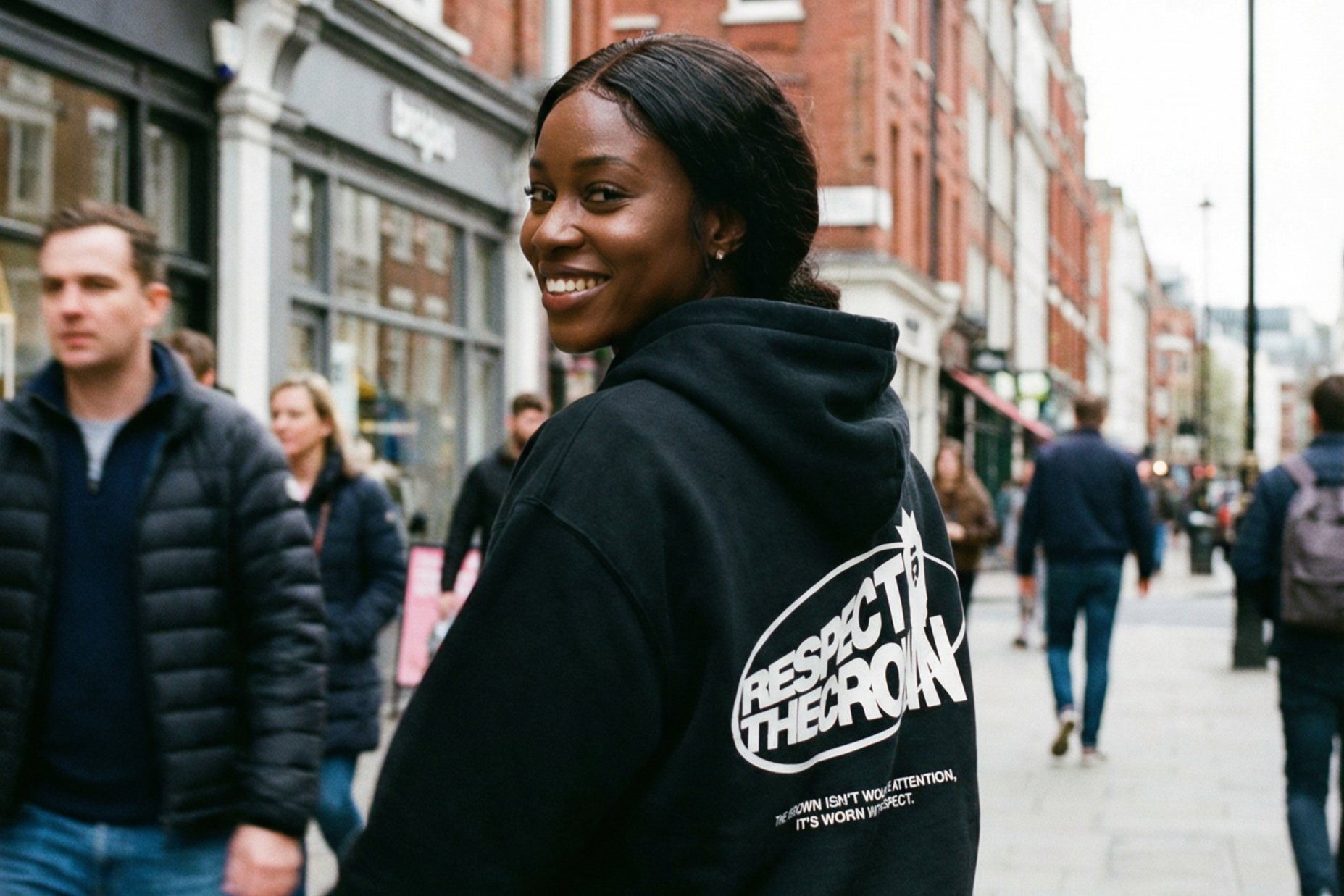 A smiling woman with dark hair, wearing a black hoodie with a slogan, stands on a busy city street with brick buildings and pedestrians.