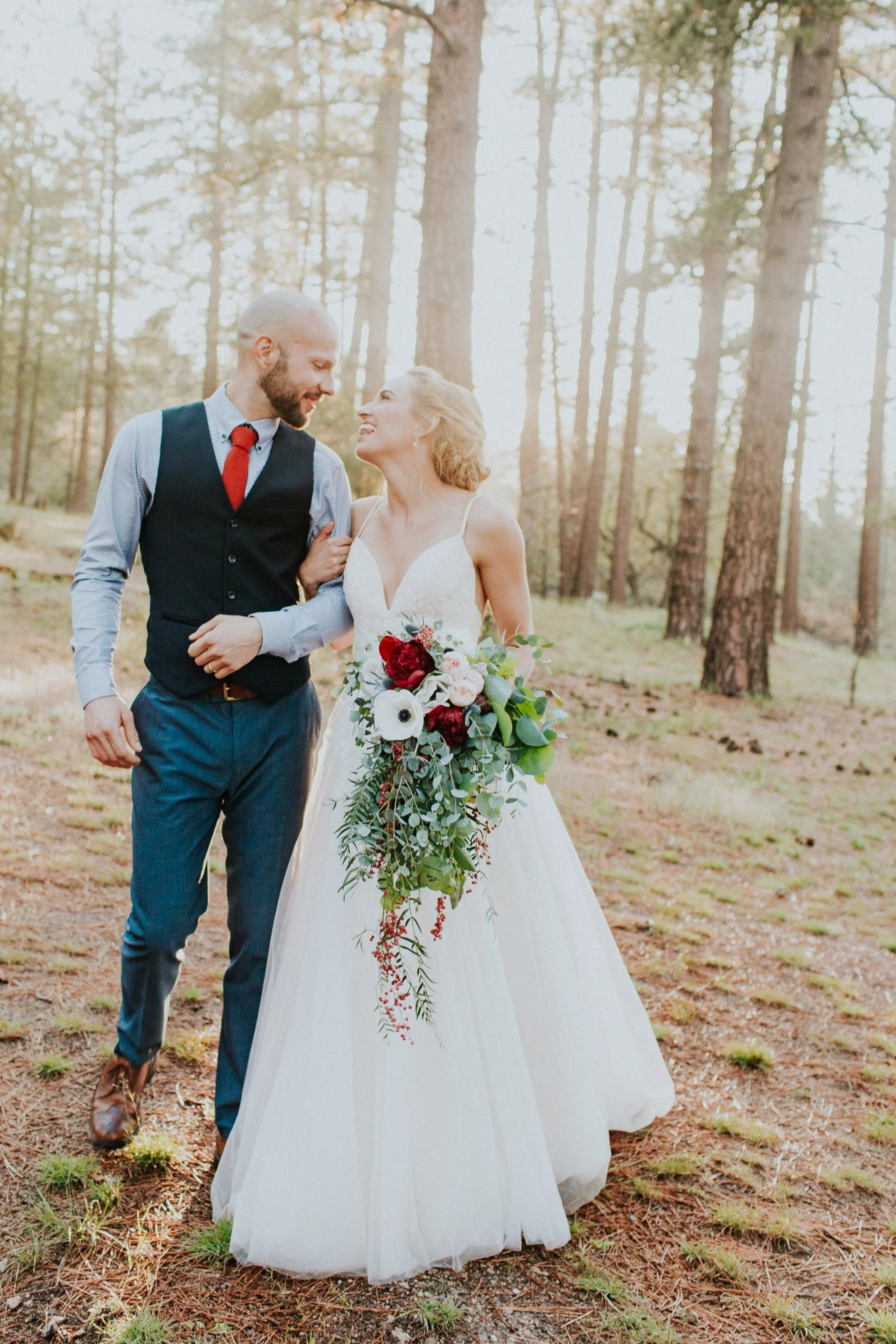 A newlywed couple walking in a forest, smiling at each other. The bride is holding a large bouquet of flowers including white anemones and red blooms. Sunlight filters through tall trees, creating a soft, romantic atmosphere.