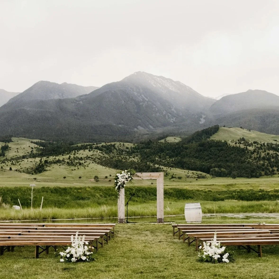 Outdoor wedding ceremony setup with wooden benches and floral arrangements in front of a mountain landscape.