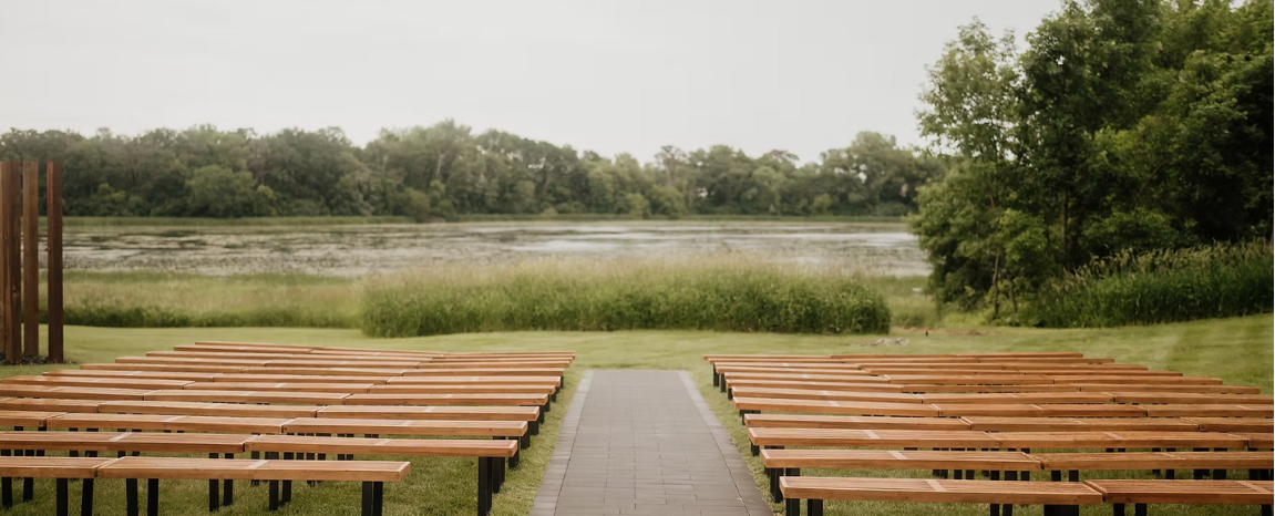 Empty wooden benches facing a lake with trees and grassy area in the background.