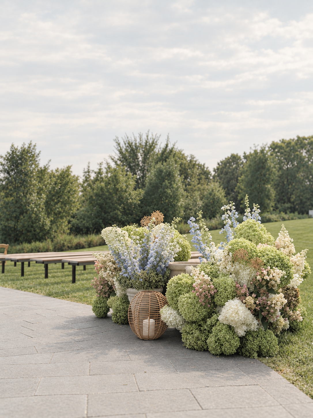 Flower arrangements with hydrangeas and delphiniums in white, pink, and blue, along with a wicker candle holder on a stone-paved outdoor area, with a grassy field and trees in the background.
