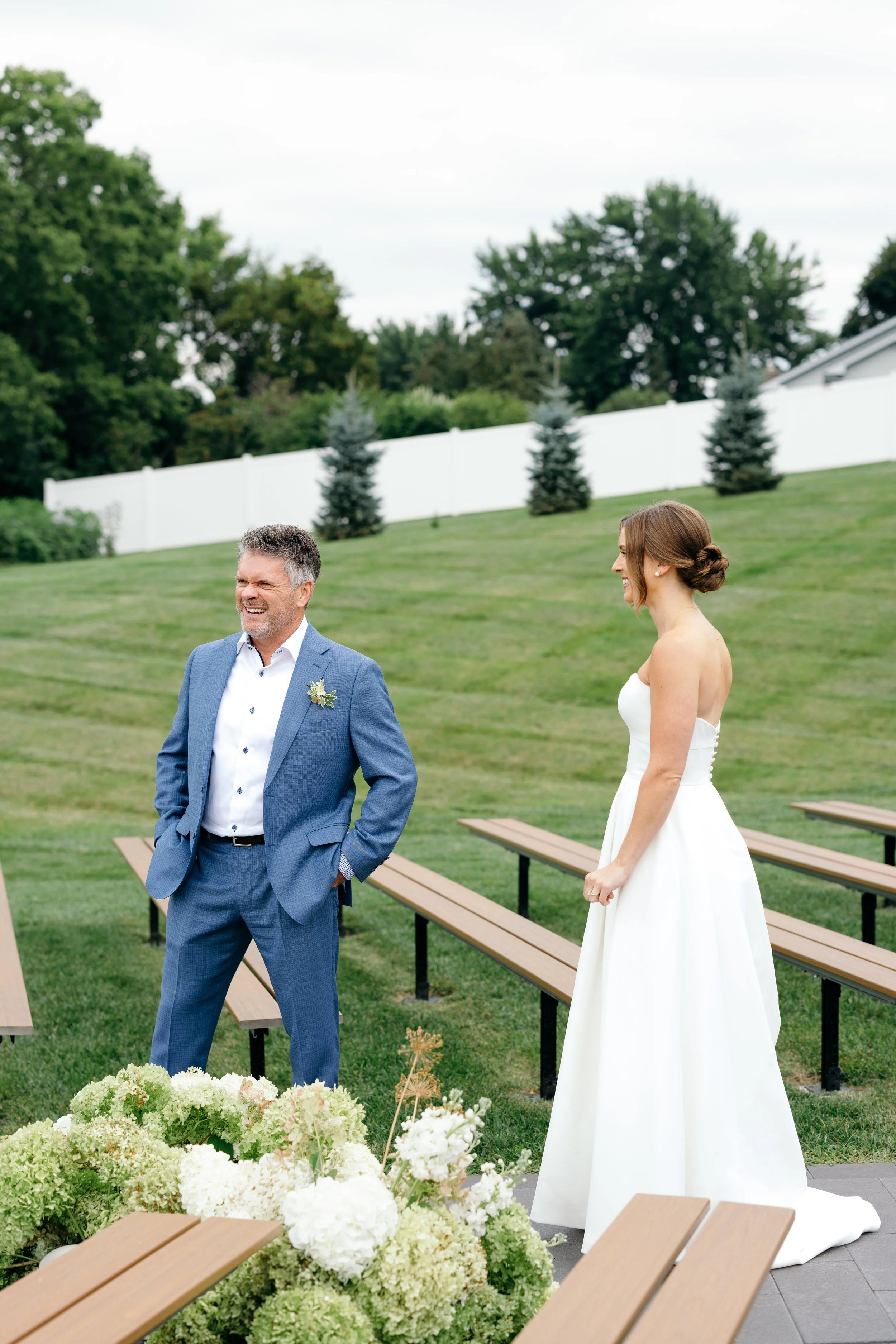A bride in a white wedding dress smiling and standing outdoors next to her father in a blue suit, during their first look with a green backyard with white fence and trees.