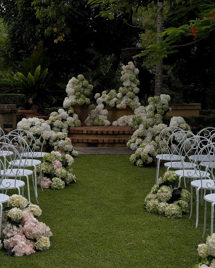 A decorated outdoor wedding aisle with white chairs on either side, white and pink floral arrangements along the aisle, and a floral backdrop at the altar with large white hydrangeas.