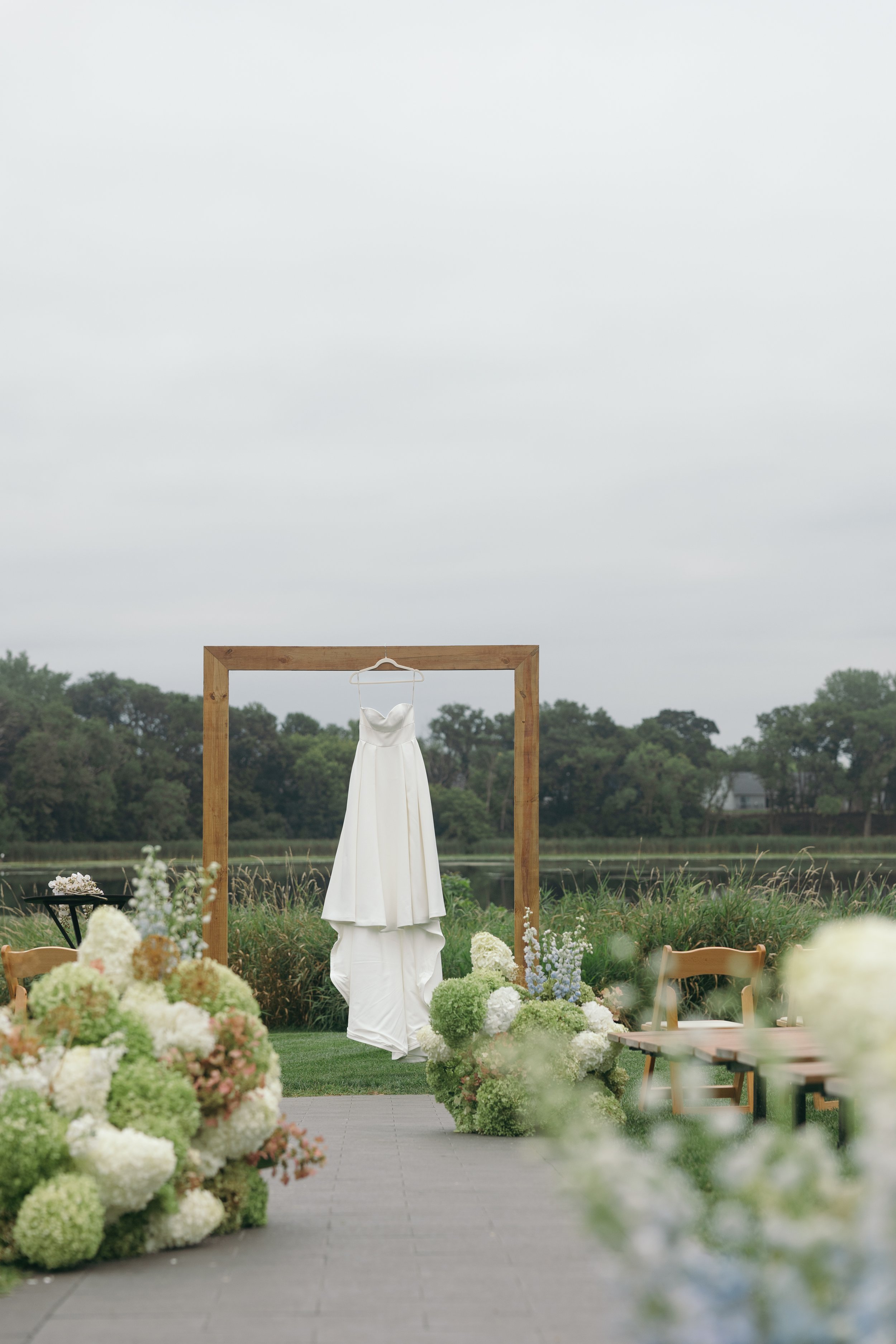 Outdoor wedding setup with a white dress hanging on a wooden frame, surrounded by flowers and chairs, with a body of water and trees in the background.