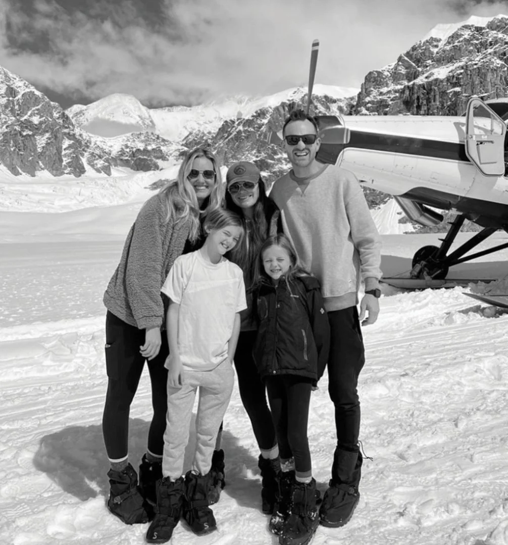 Family photo of five people standing on snow with mom, dad, kids smiling in front of plane on mountain in Alaska.