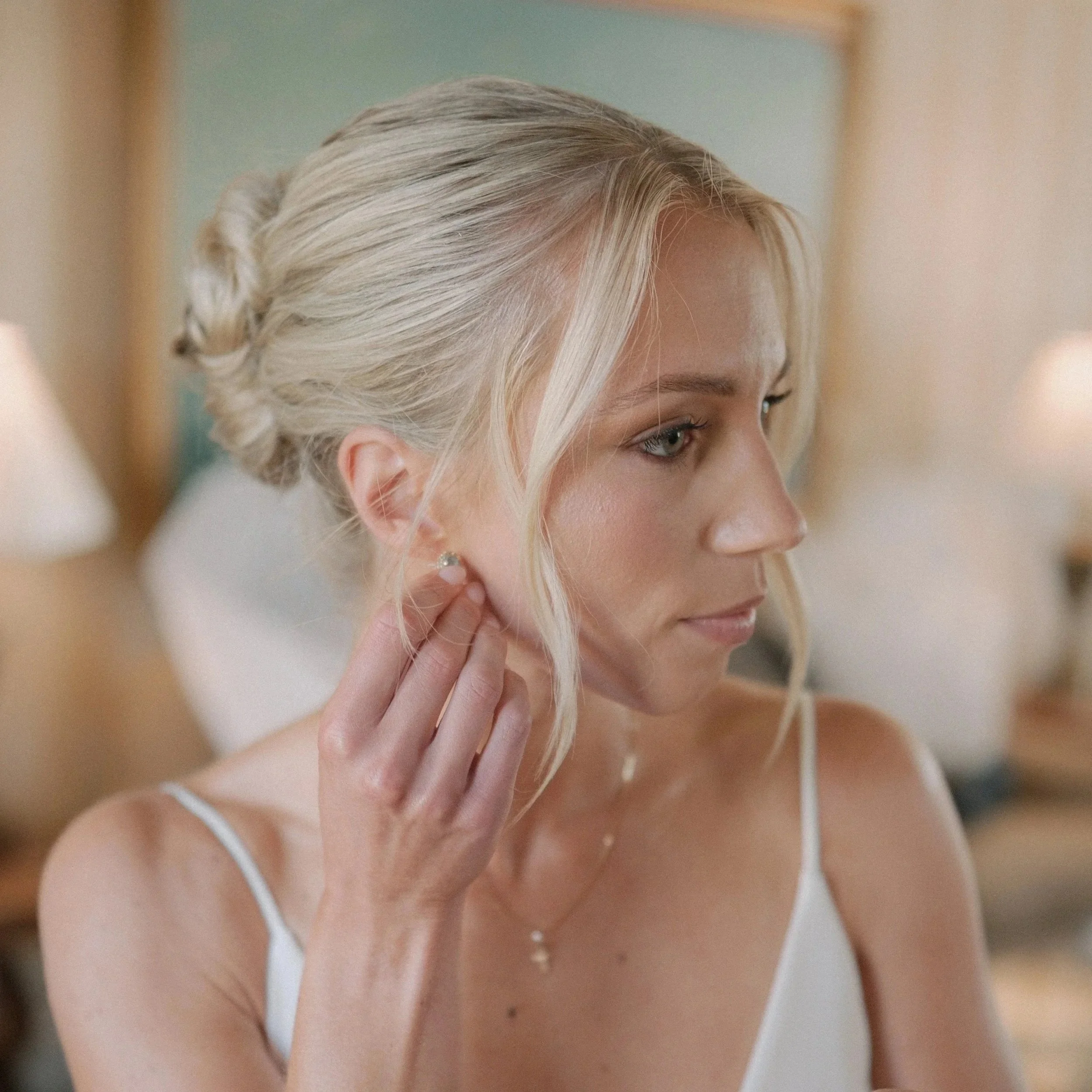 A woman with blonde hair styled in an updo, putting on earrings in a softly lit bedroom.