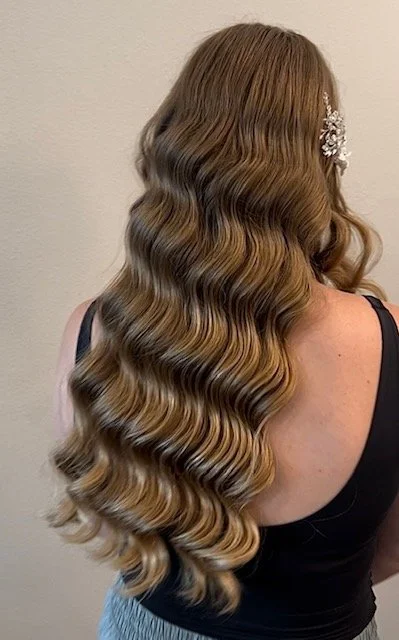 Back view of a woman with long, wavy, honey-blonde hair wearing a black top and a decorative hair accessory.