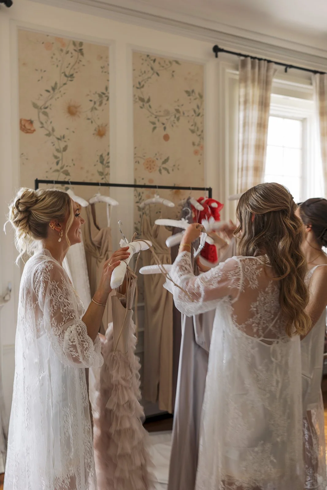 Women in lace robes hanging Christmas stockings on a clothing rack in a bright, floral-patterned room.