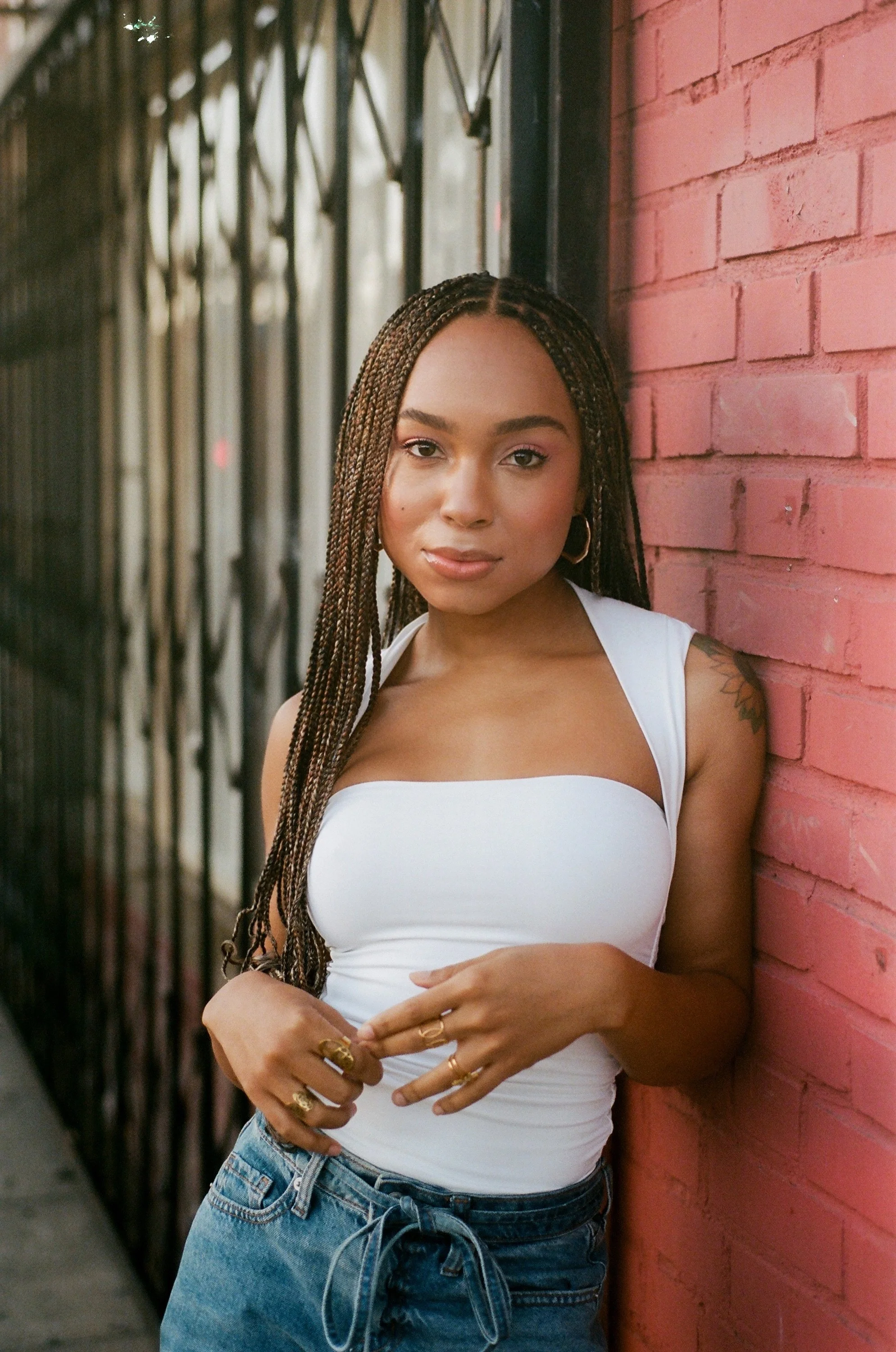 Young woman with braided hair and hoop earrings standing by a pink brick wall, wearing a white top and blue jeans.