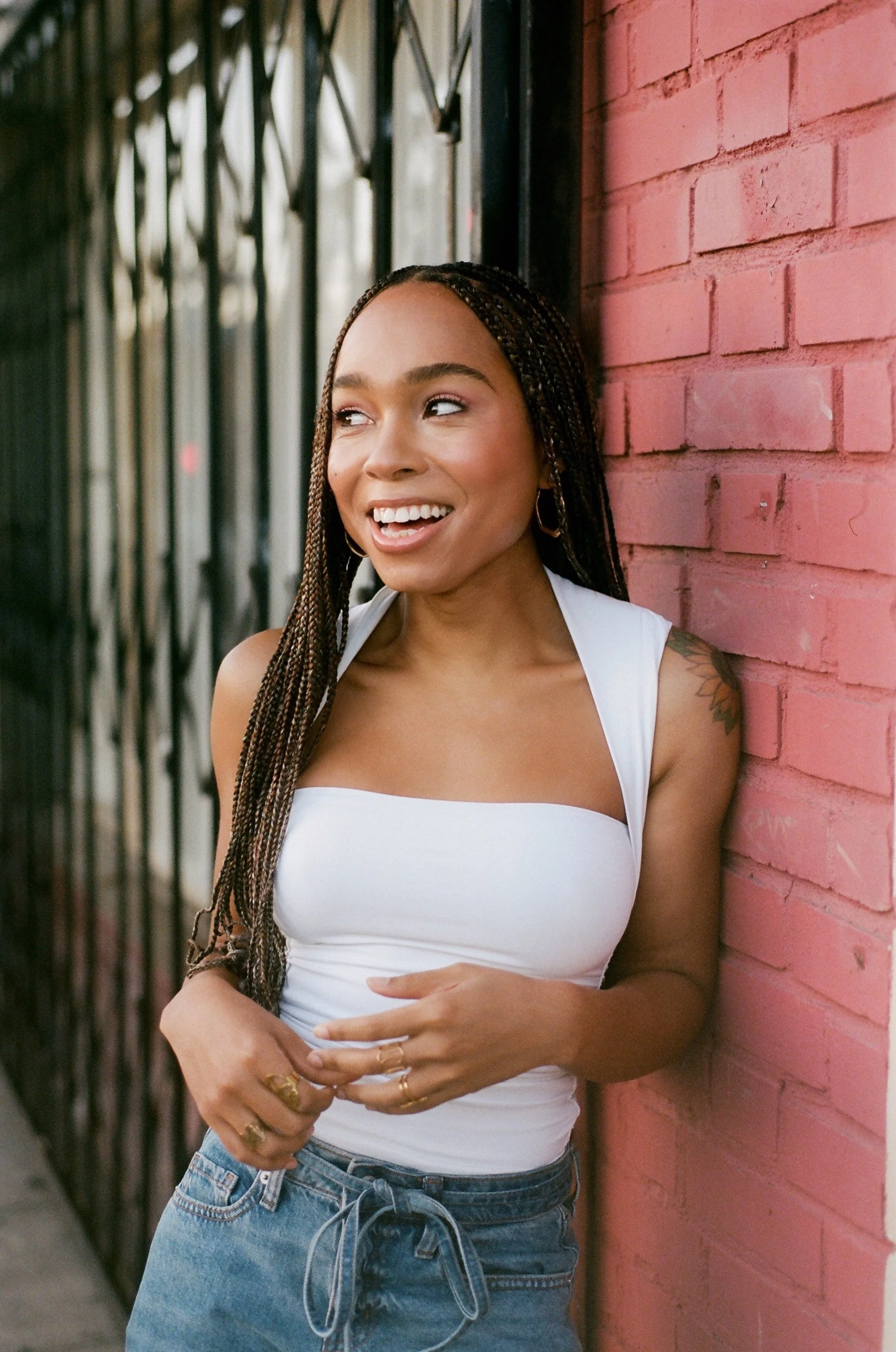 A young woman with long braided hair wearing a white tank top and denim shorts, leaning against a red brick wall, smiling and looking to the side.