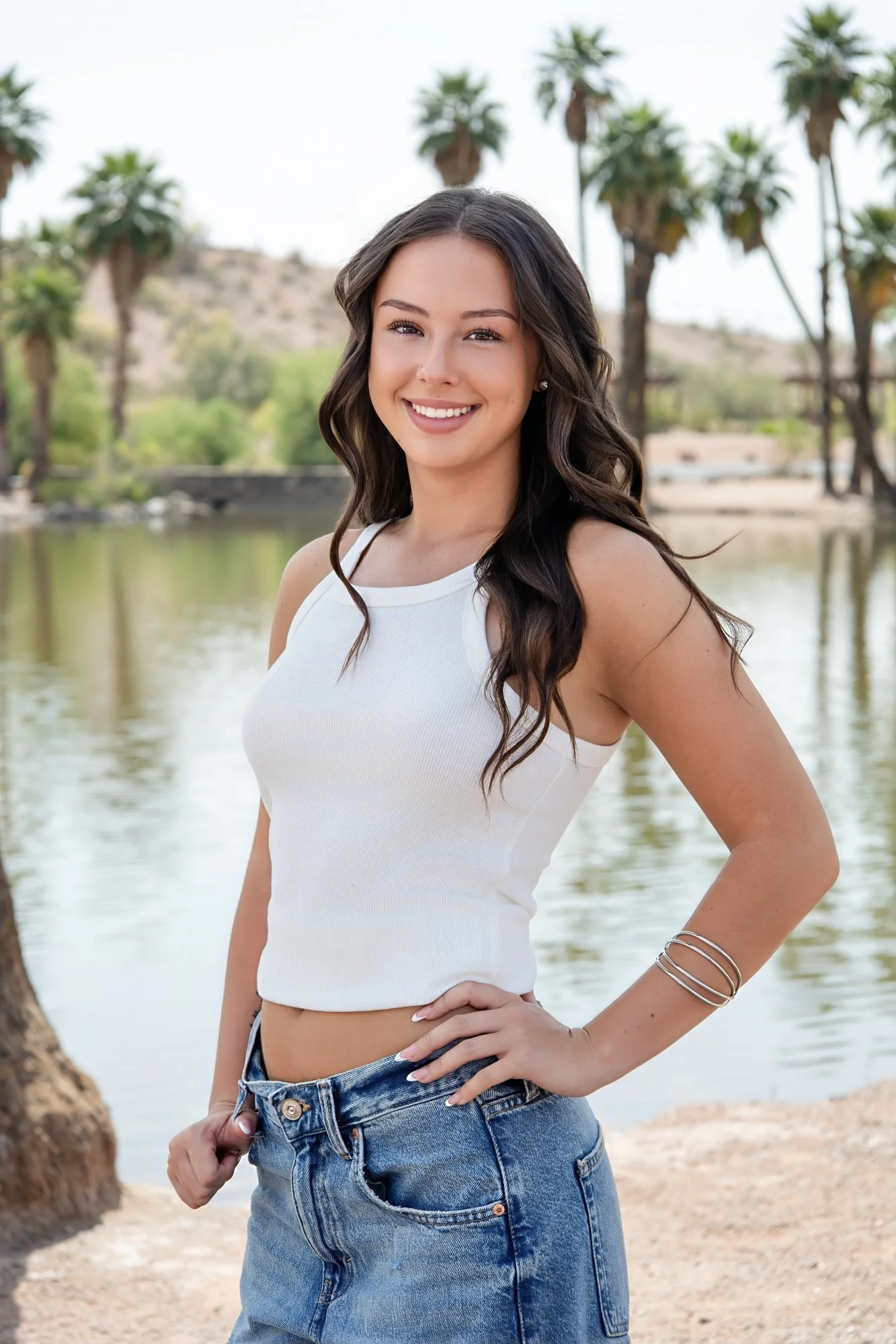 Senior portrait: girl with long dark hair in a white tank top and jeans by a pond with palm trees