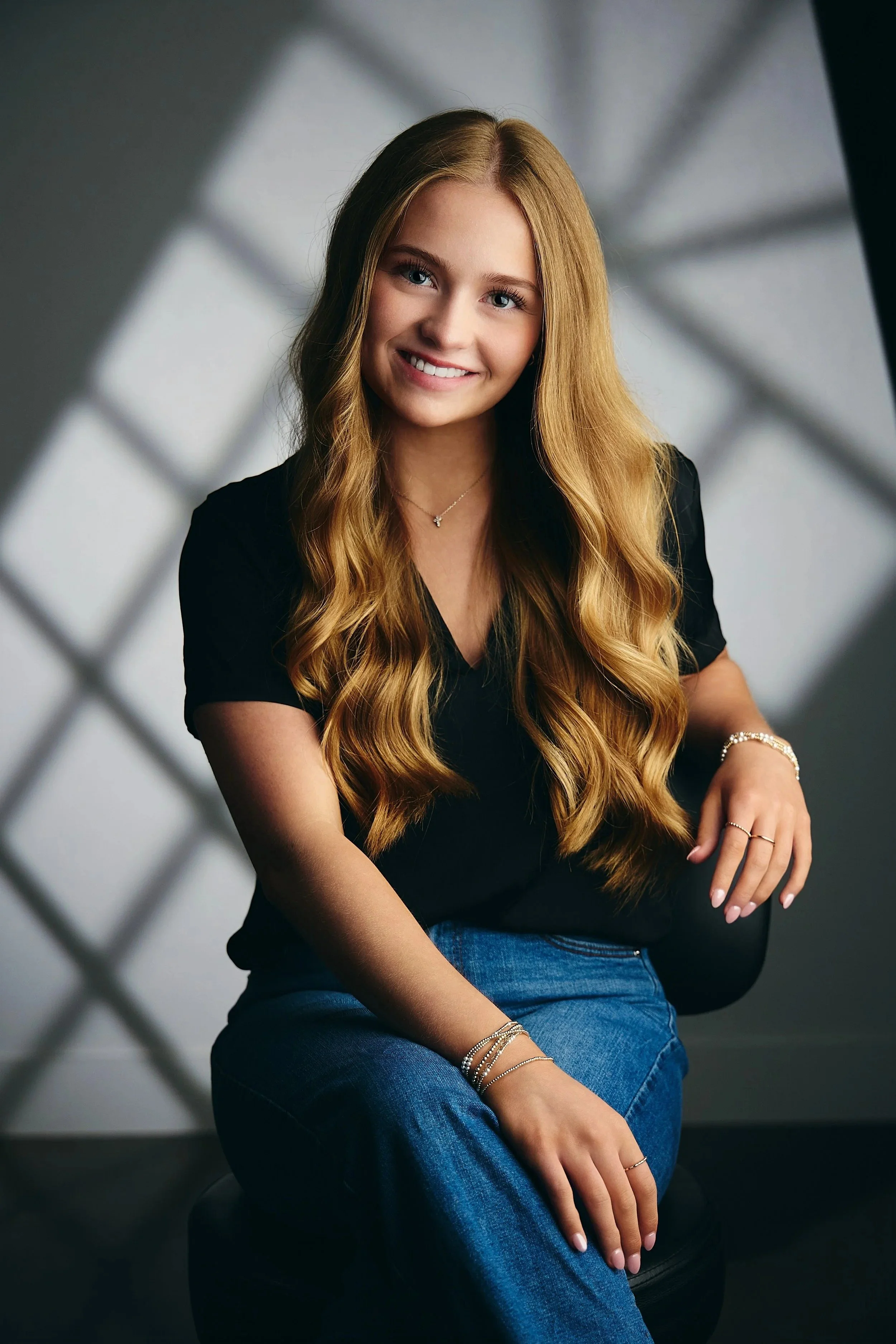 Seated senior picture pose: girl with long blonde hair in a black top and blue jeans on a black chair