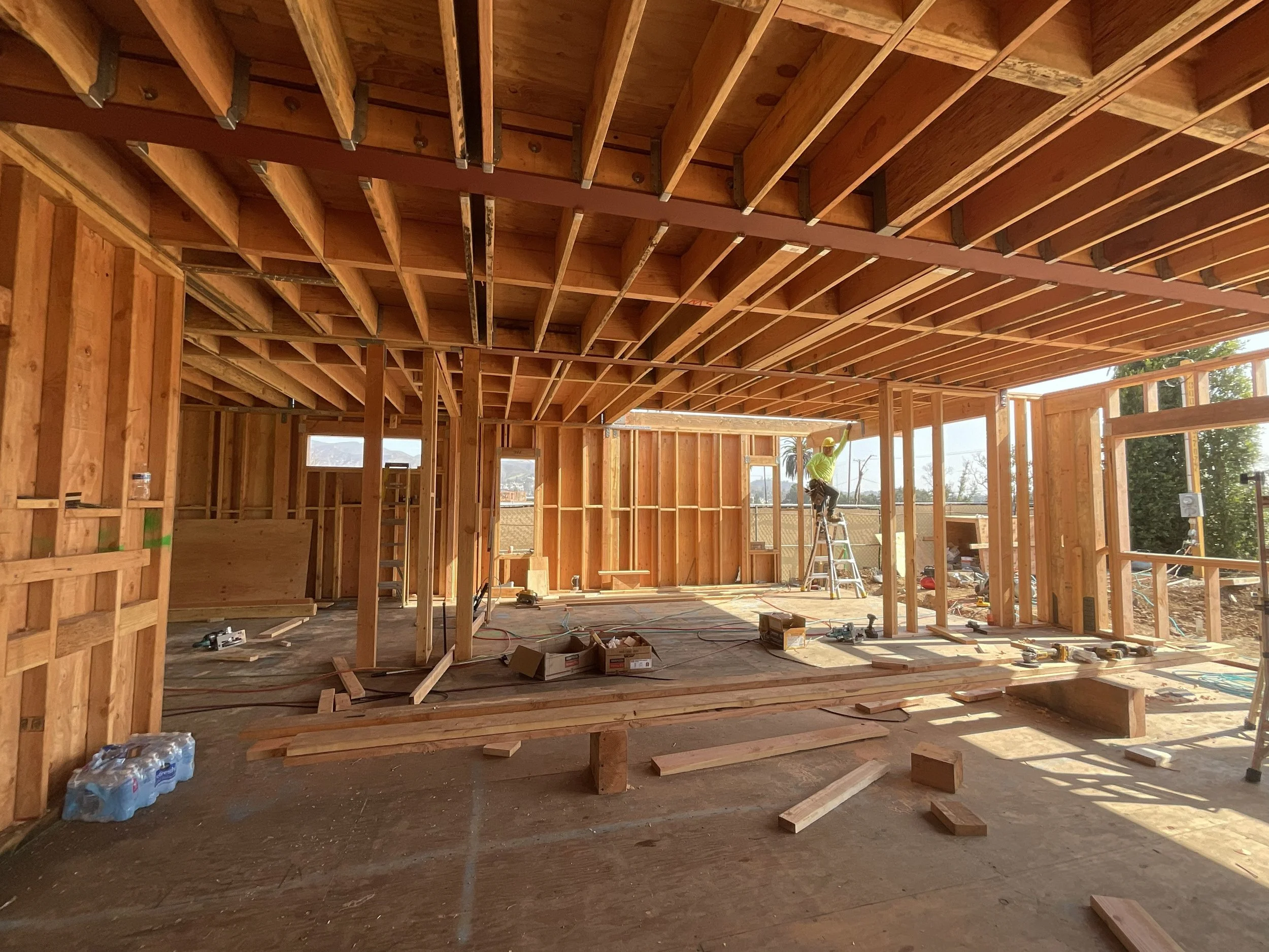 Interior view of a house under construction with exposed wooden framing, some walls, and a worker on a ladder.