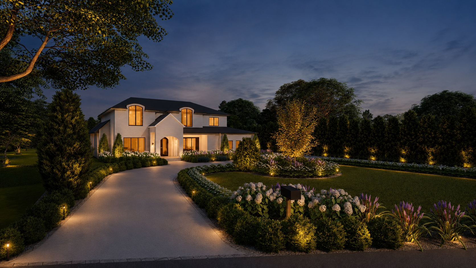 A large, modern house is illuminated at dusk with warm lights, surrounded by a landscaped garden with trees, bushes, and flowering plants, and a curved pathway leading to the entrance.