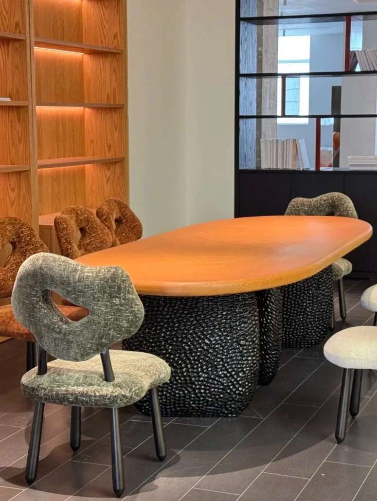 A modern conference room with a wooden oval table, six chairs with varied upholstery, and a background consisting of a wooden bookshelf and partition with glass panels.