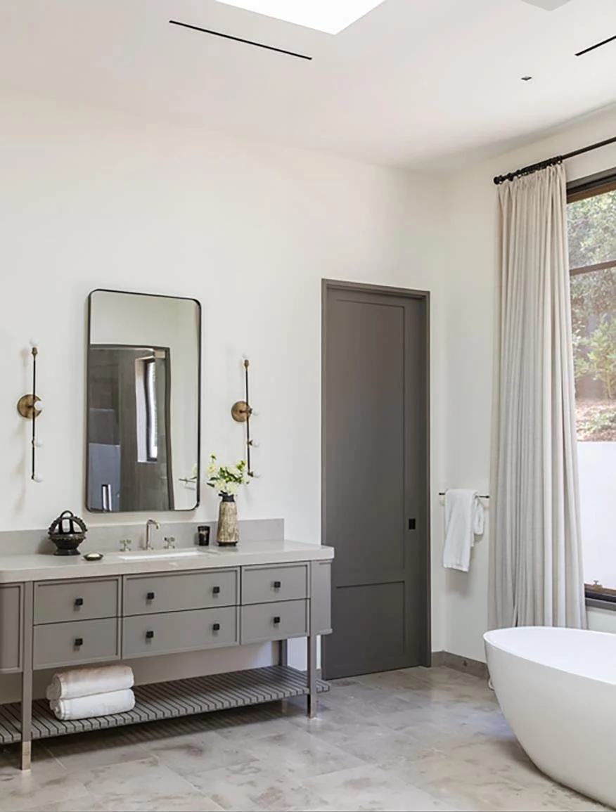 Modern bathroom with a white bathtub, gray vanity with a mirror, towels, a vase with flowers, and a window with white curtains.