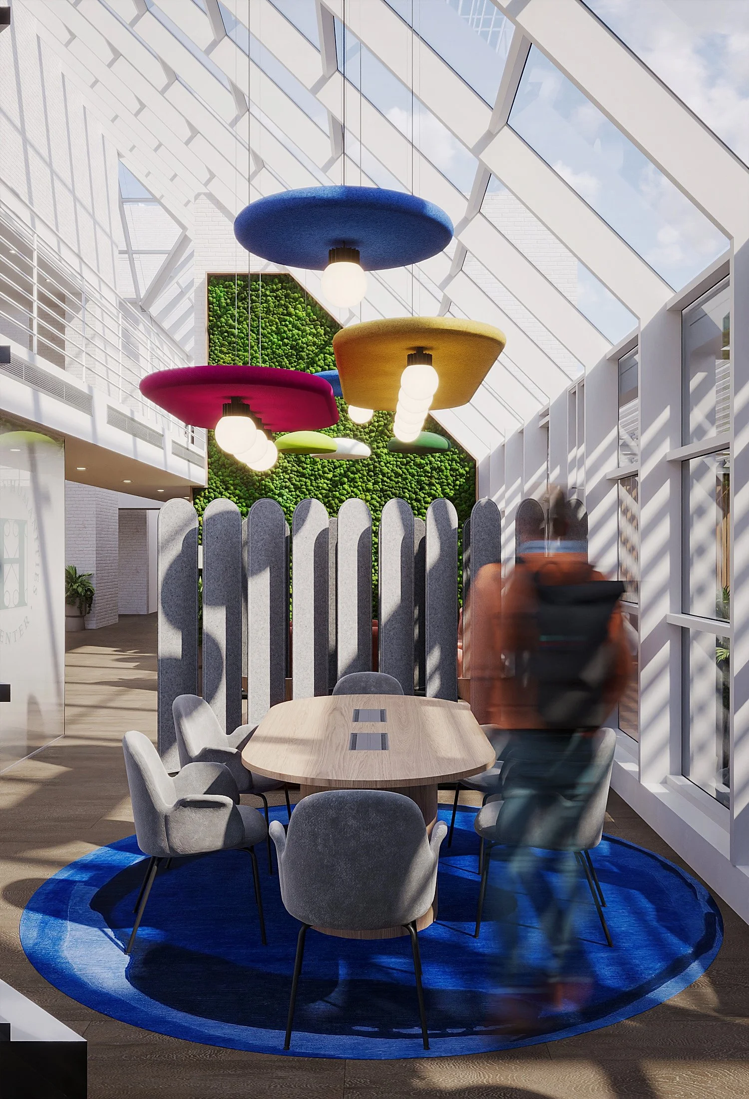 Modern interior space with a round wooden dining table surrounded by gray upholstered chairs on a blue circular rug. Overhead, colorful hanging lamps in blue, yellow, pink, and green. The background features a green vertical garden wall and a ceiling with large glass panels allowing natural light and a view of the sky.