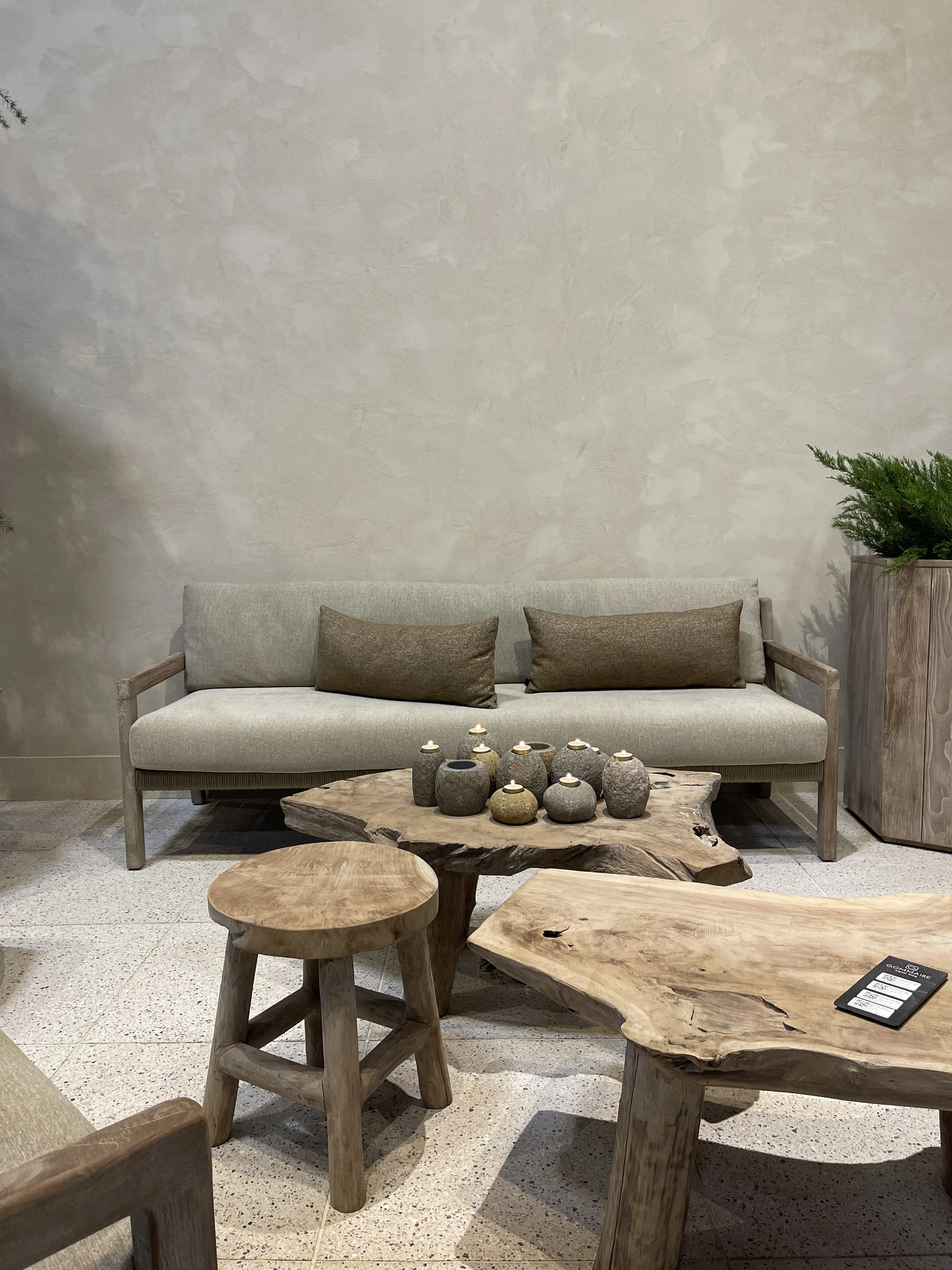 Outdoor furnitures with a light-colored sofa with two brown pillows, a rustic wooden coffee table with candles, a matching wooden side table, and a potted plant on the right, against a textured beige wall.