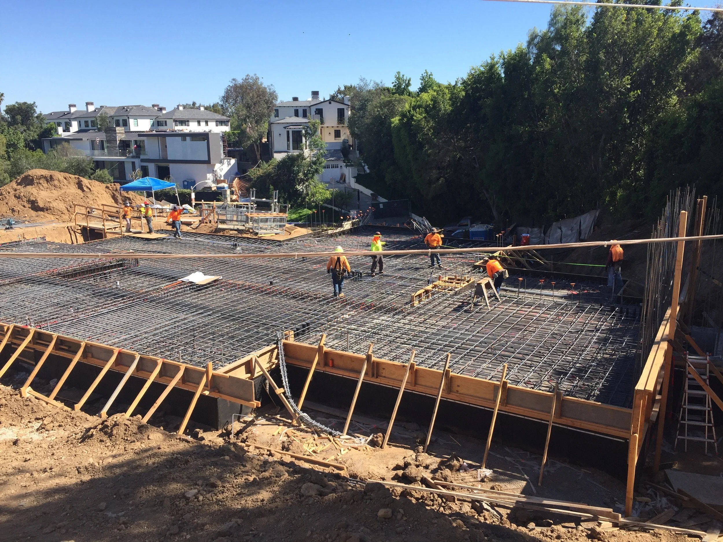 Construction site in Beverly Hills with workers building a concrete foundation, surrounded by wooden framing and rebar, with residential houses and trees in the background.