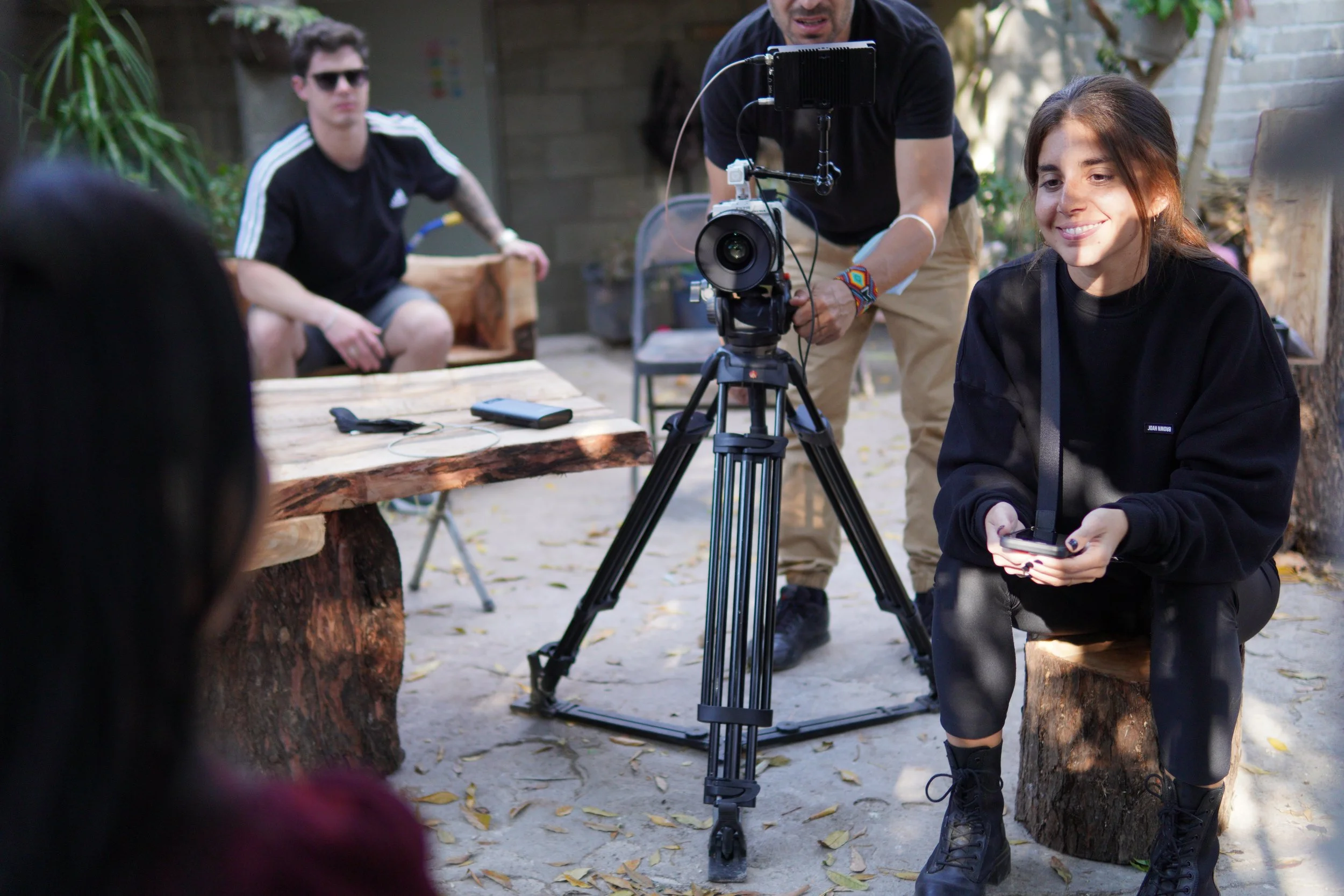 A woman kneels next to a tripod-mounted camera setting up a shot outdoors, with a man adjusting the camera and another person sitting on a log in the background.