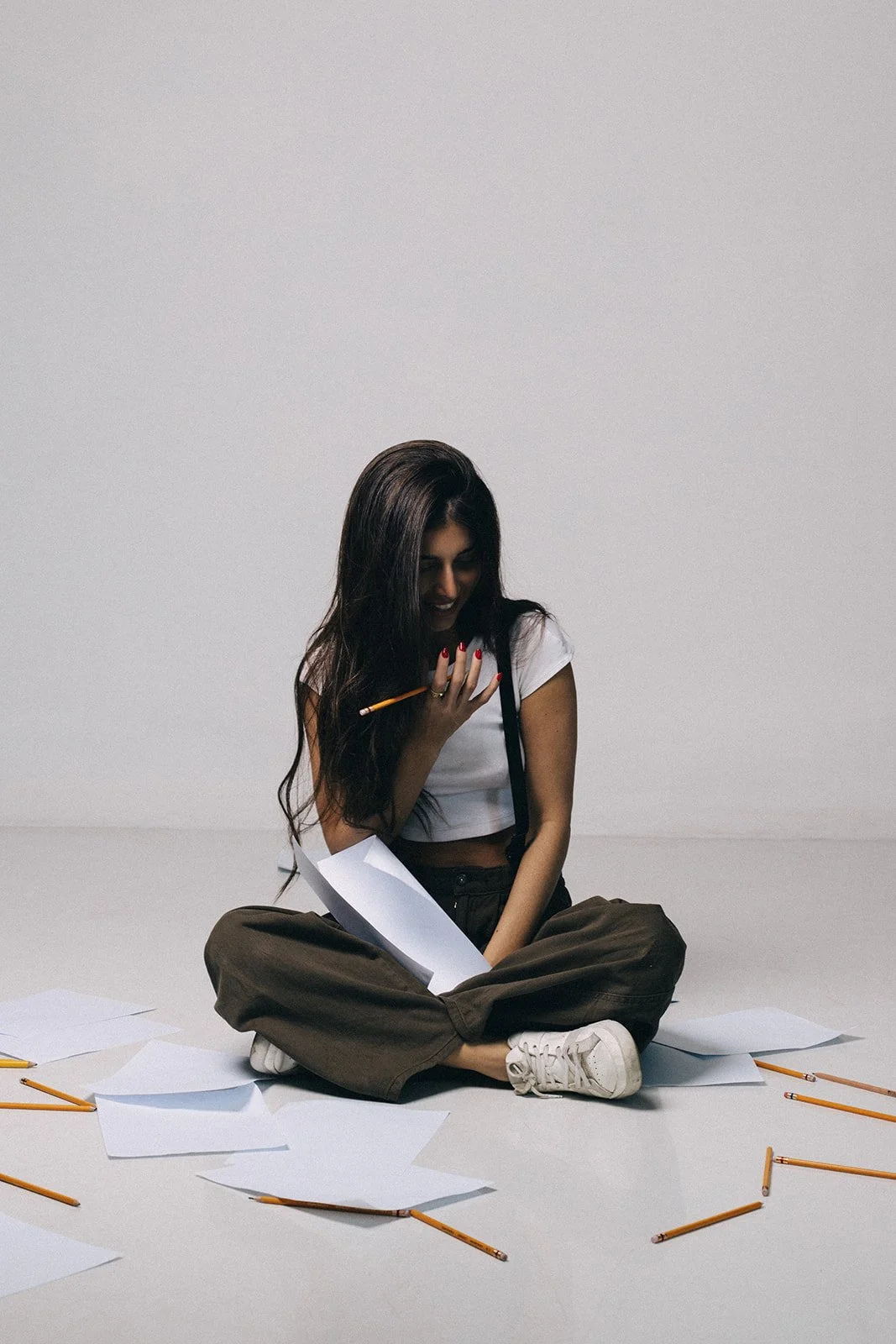 Young woman sitting cross-legged on the floor with scattered papers and pencils, smiling and holding a pencil near her face, against a plain white background.