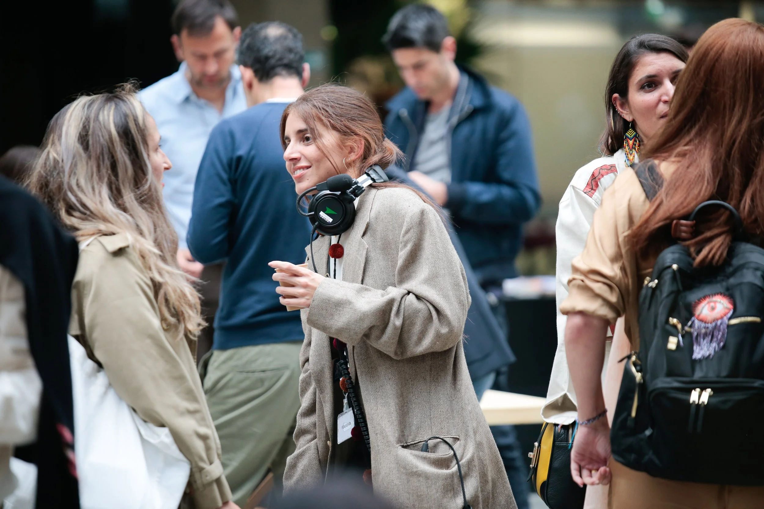 Group of people outdoors, engaged in conversation, including a woman wearing headphones and a beige blazer smiling with others around her.