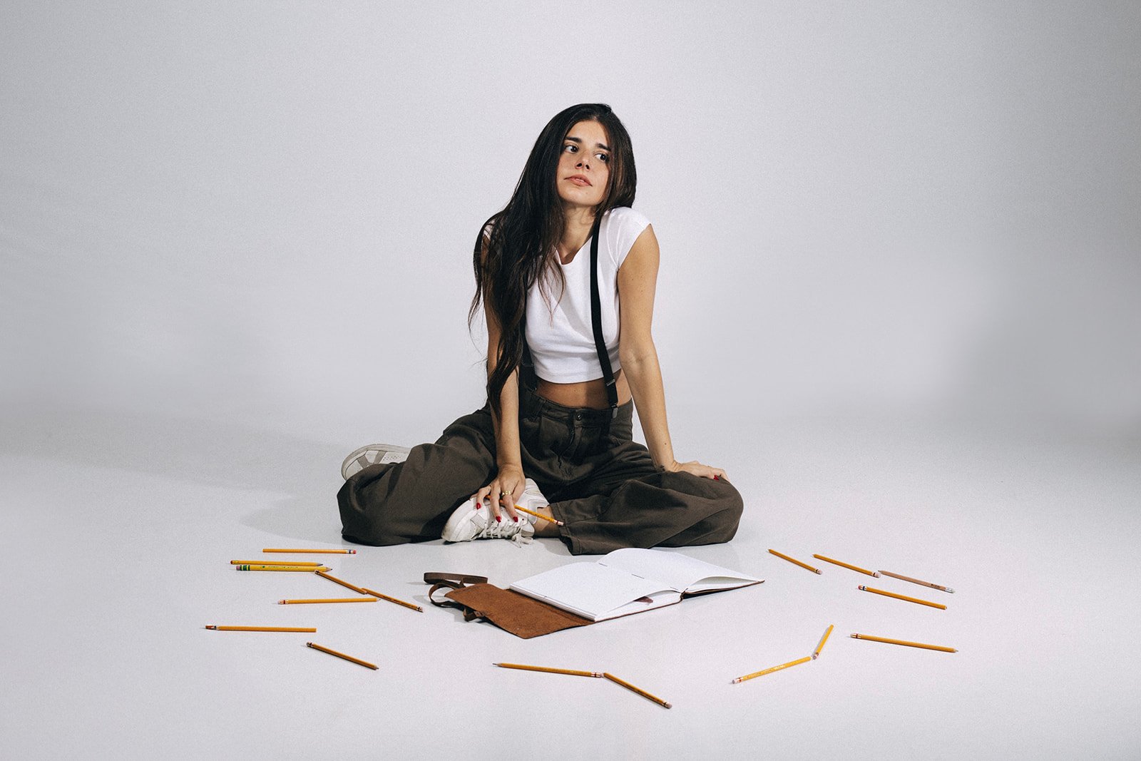 A young woman with long dark hair sitting cross-legged on the floor in front of an open book and scattered pencils, looking contemplative.