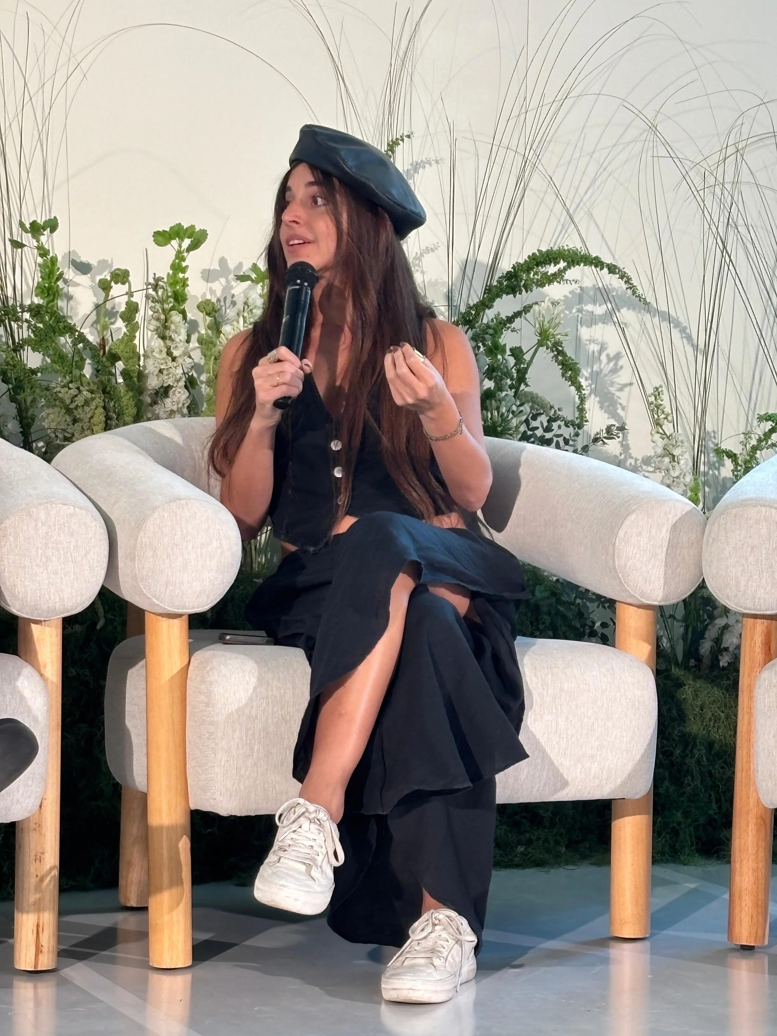 A woman with long brown hair, wearing a black leather beret, a black sleeveless top, and black pants, kneeling on one knee, holding a microphone, in front of a white wall with tall green plants. She is wearing white sneakers and sitting on a beige sofa with wooden legs.
