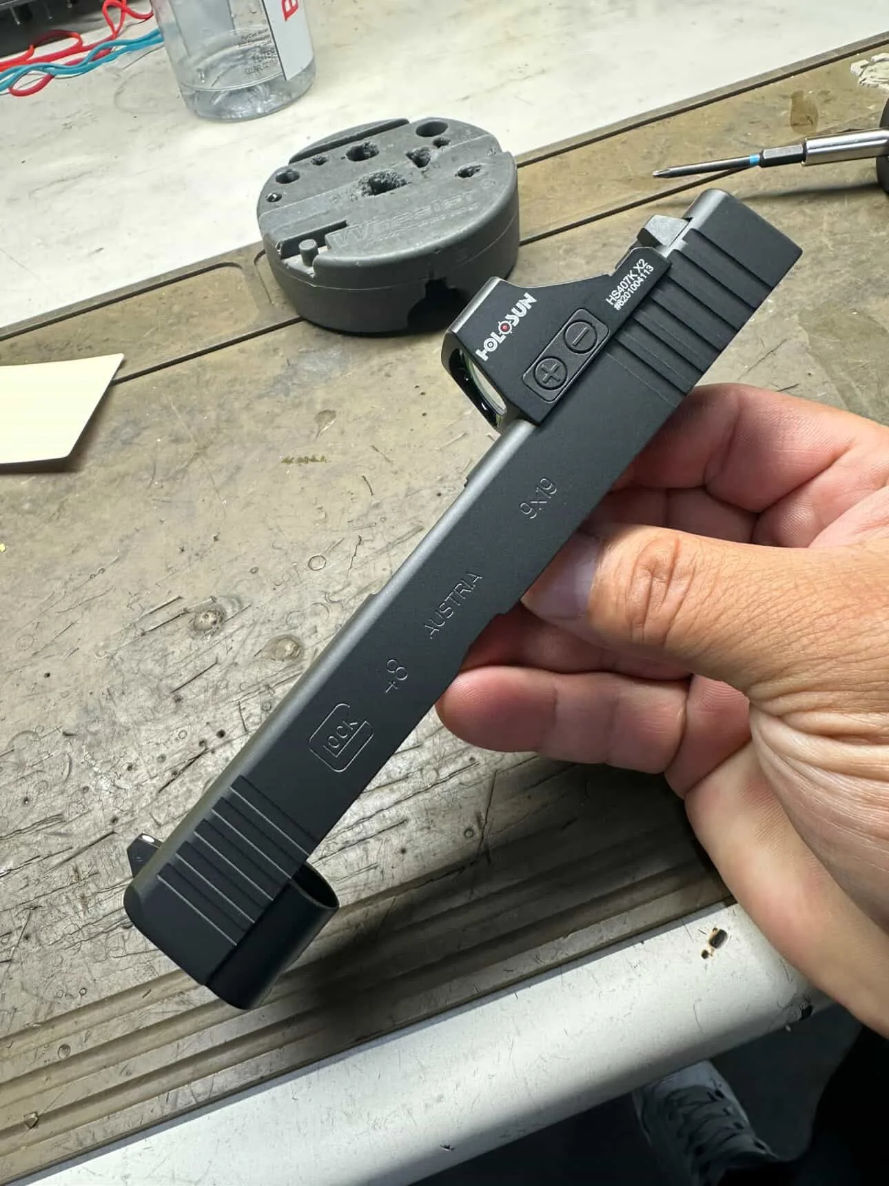 Custom Glock 48 pistol slide with a red dot sight attached, on a wooden workbench with a gray component, screwdriver, and other tools.