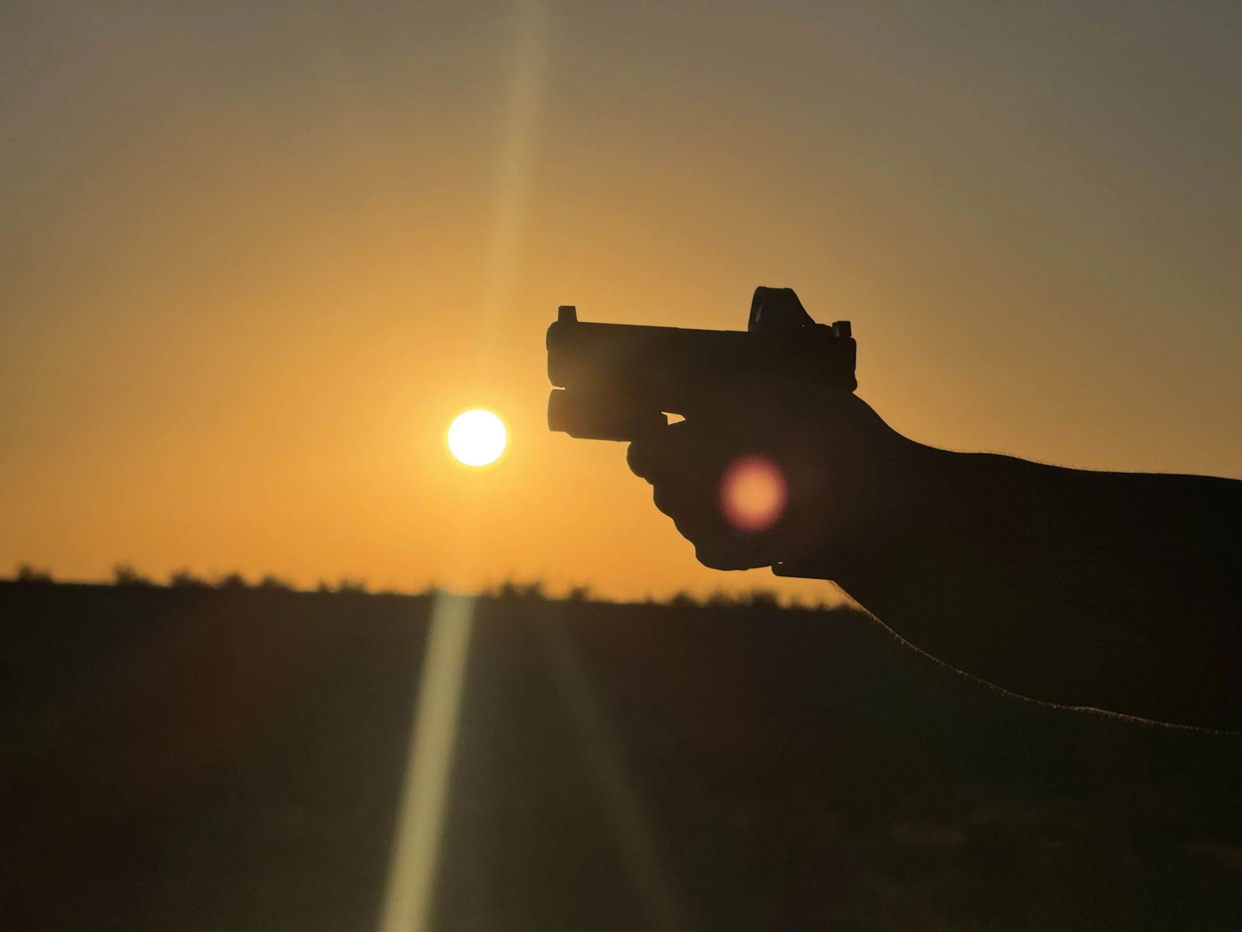 Silhouette of a hand holding a handgun against a sunset sky with the sun visible near the gun's barrel.