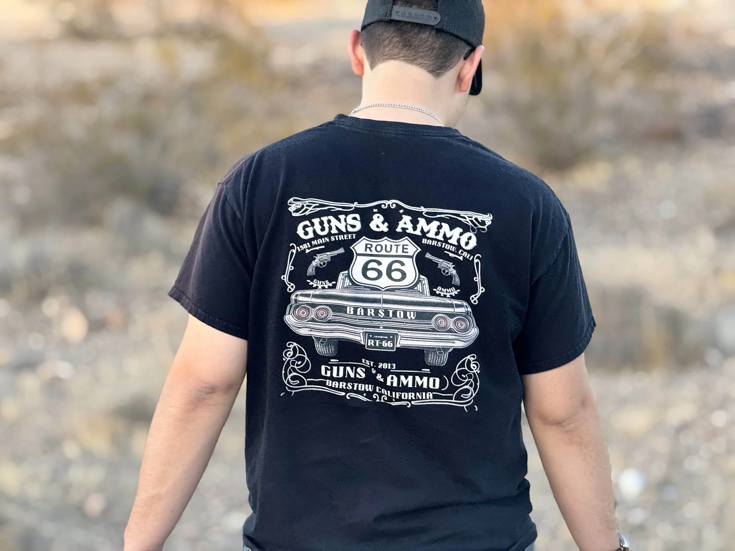 Back view of a person wearing a black t-shirt with white graphic design, including Route 66 sign, guns, and ammo text, outdoors in a desert-like area.