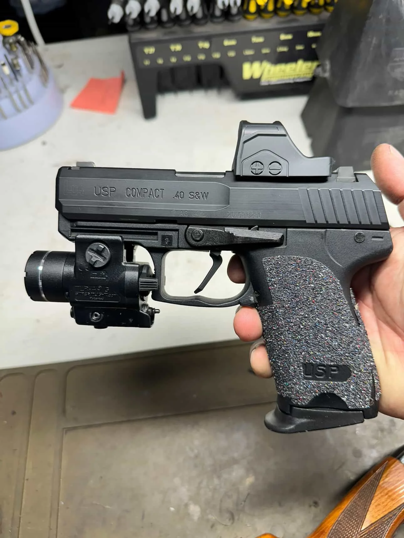 A person holding a black semi-automatic pistol with a red dot sight mounted on top, in a workshop or gun store setting.