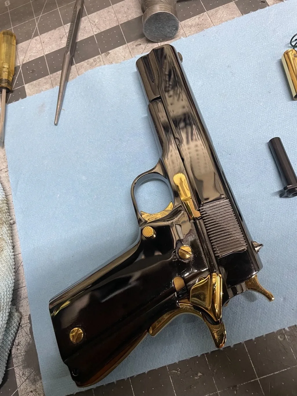 A metallic, black pistol with gold accents laid on a blue cloth surface, surrounded by tools and parts on a workbench.