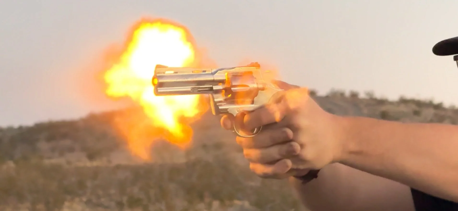 A person firing a handgun, with a bright muzzle flash and smoke coming from the barrel, outdoors on a desert landscape.
