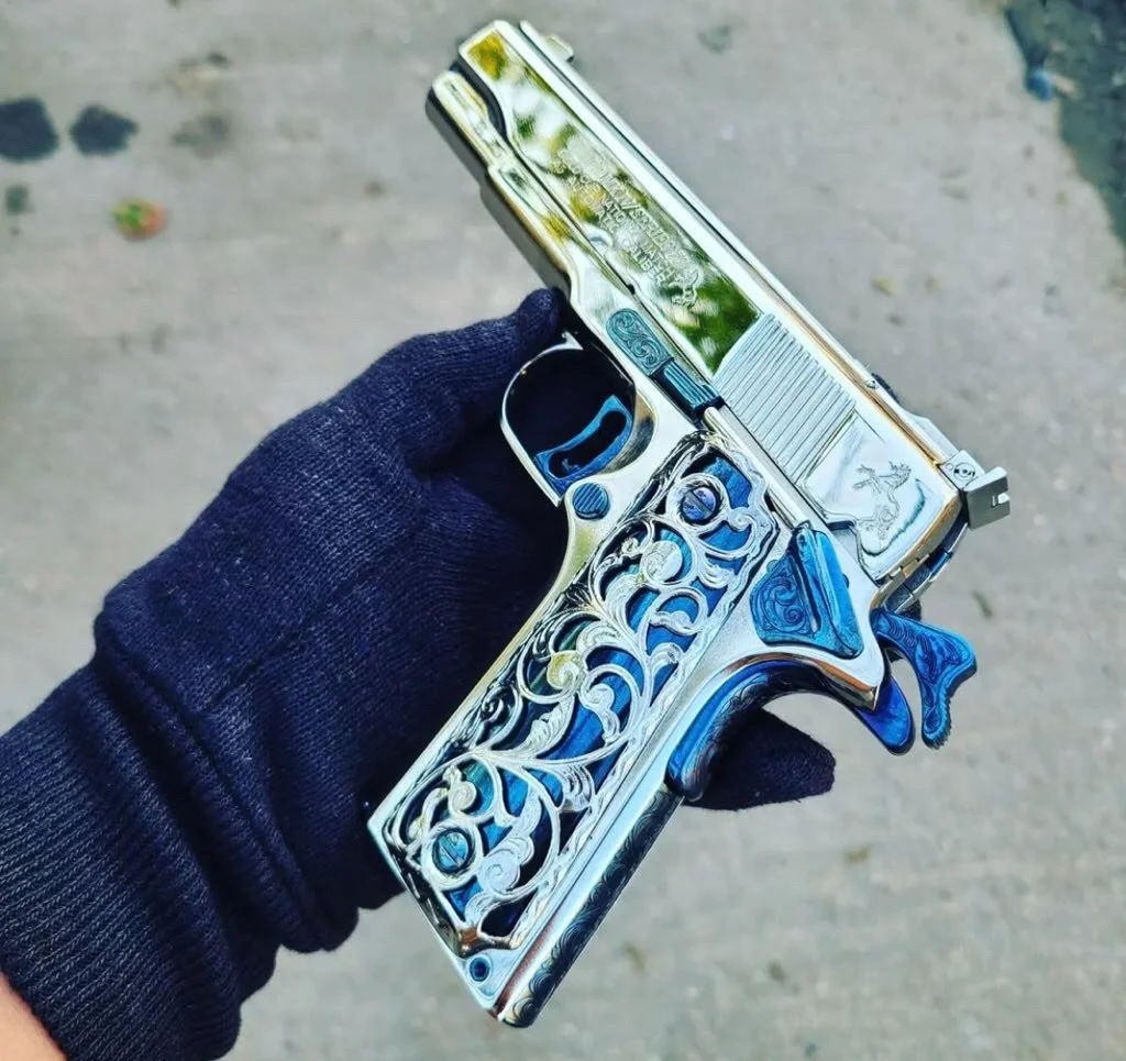 A silver handgun with intricate blue and white designs engraved on the handle and slide, being held by a person wearing a dark glove.