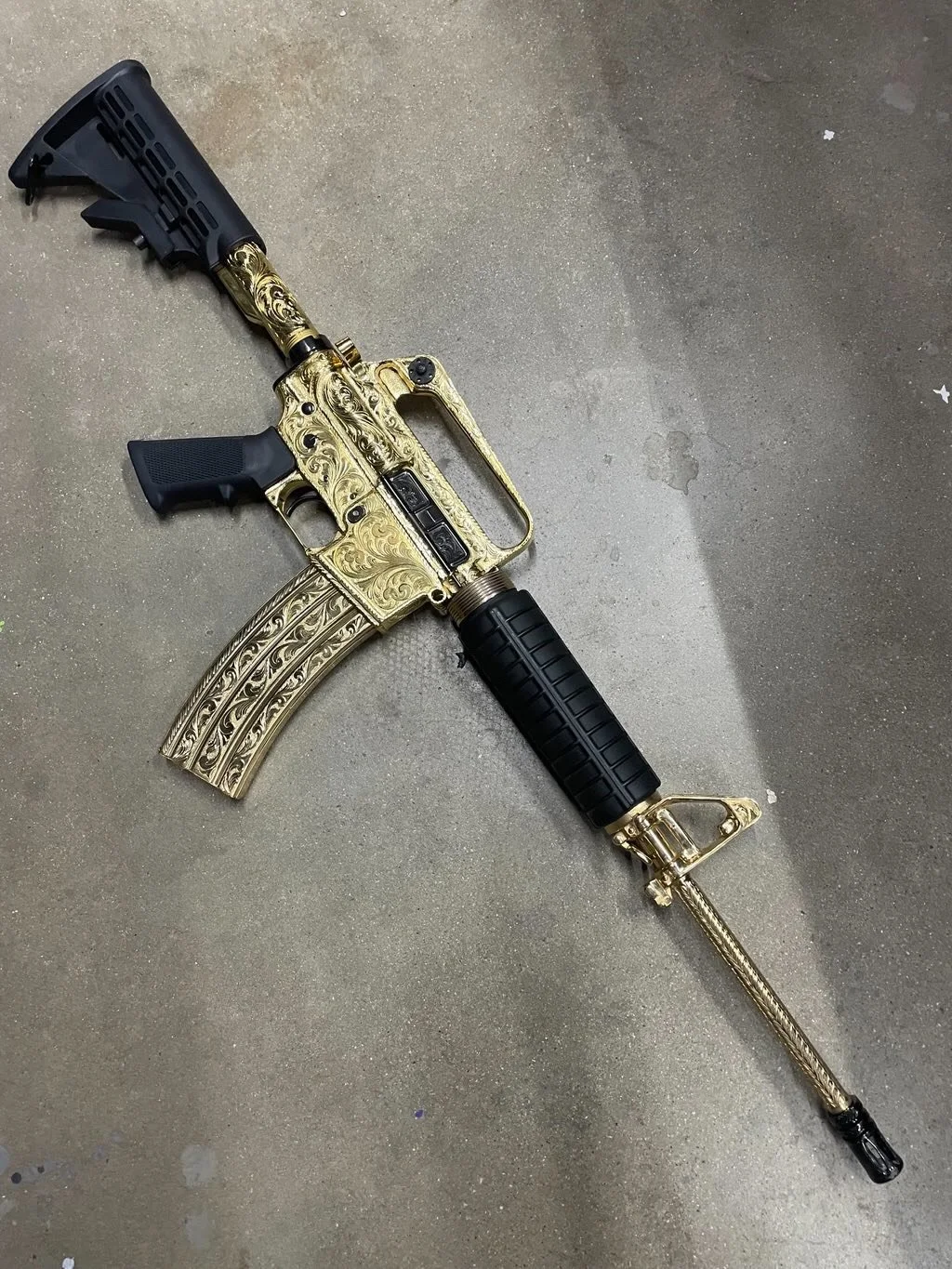 A gun with intricate gold engravings, black grip and stock, resting on a concrete surface.