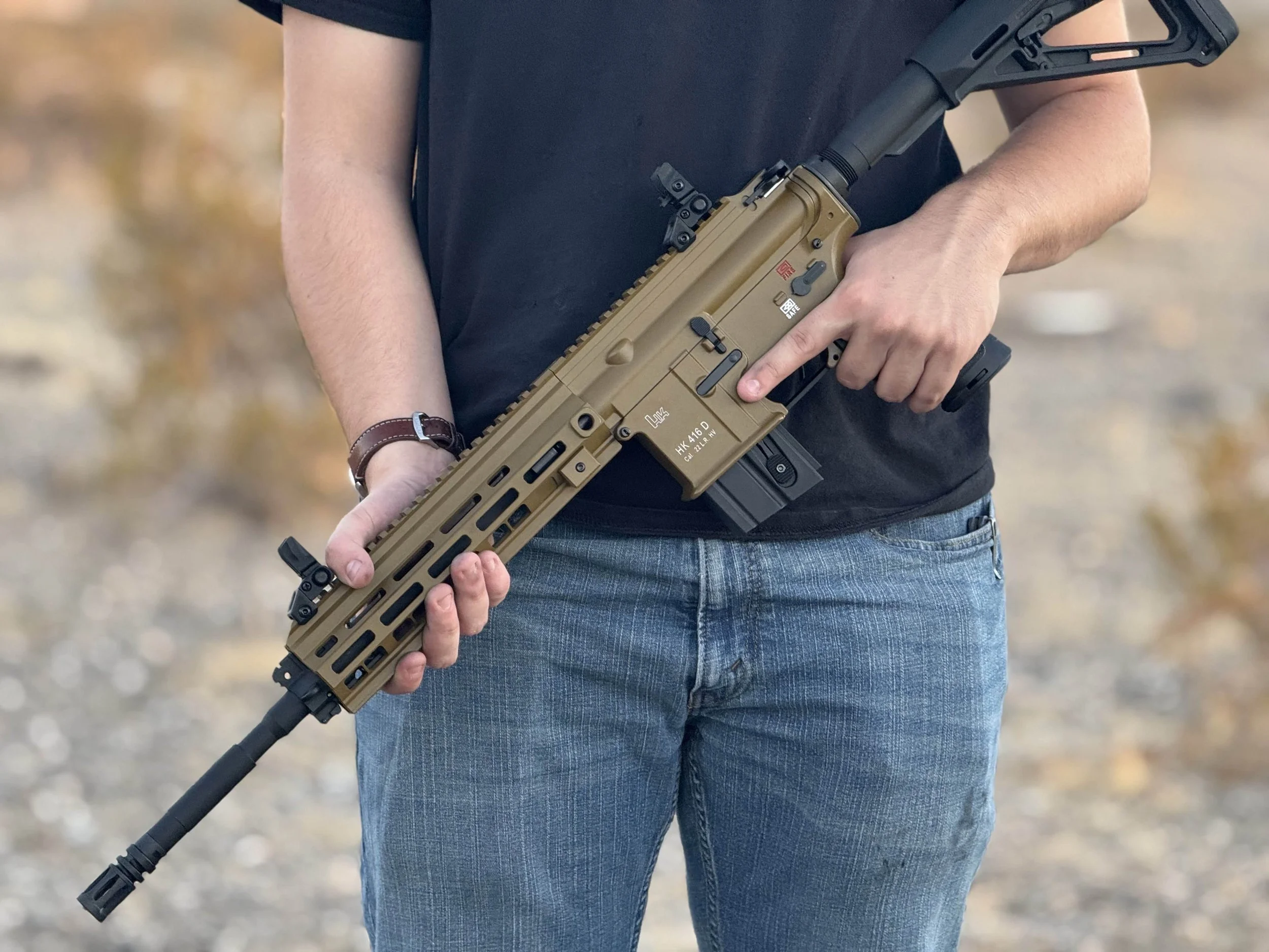 Person holding a tan-colored semi-automatic rifle with a black barrel, in outdoor setting. The person is wearing a black T-shirt, blue jeans, and a wristwatch.