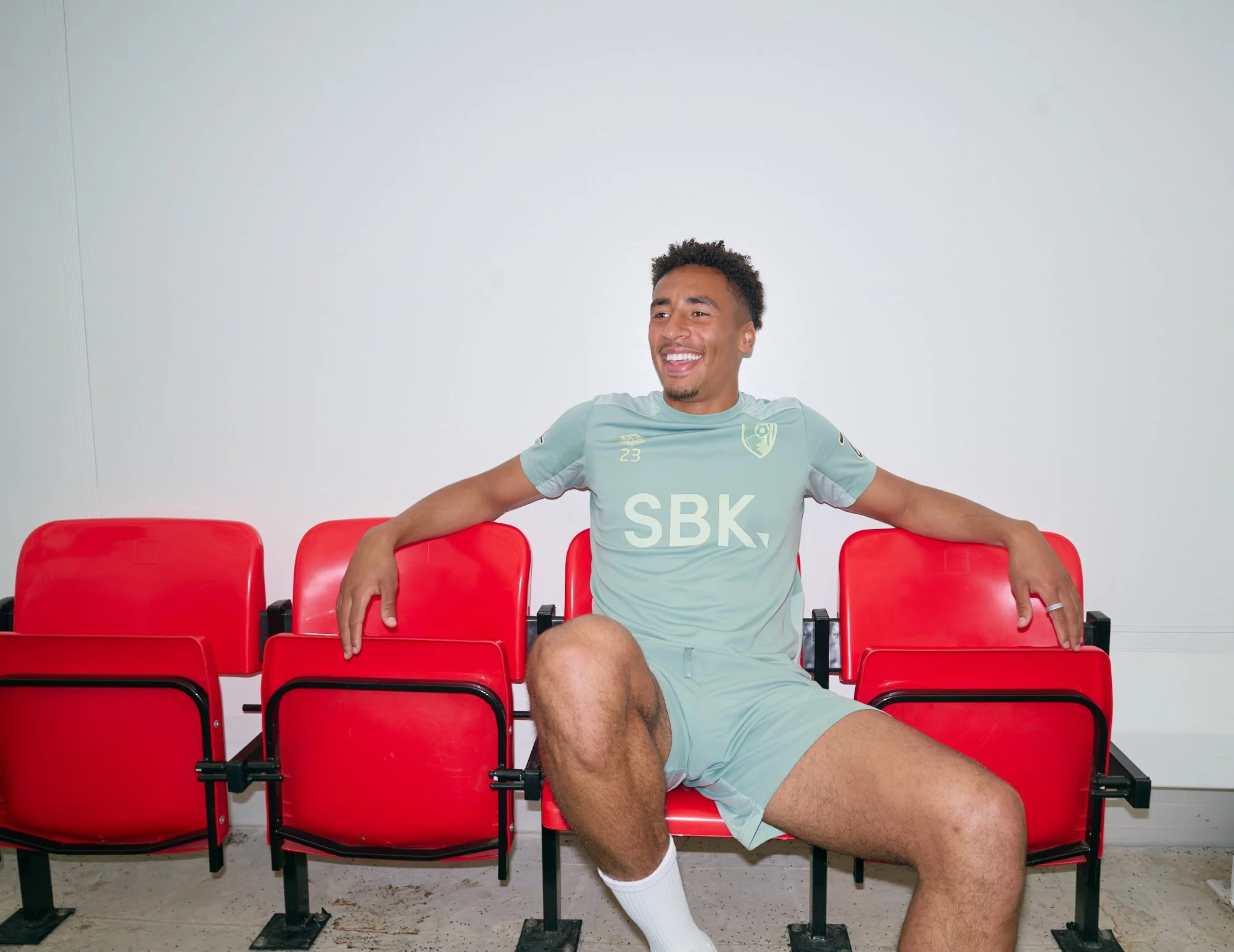 Professional footballer James Hill smiling as he sits across a row of bright red stadium seats, wearing an AFC Bournemouth training kit.