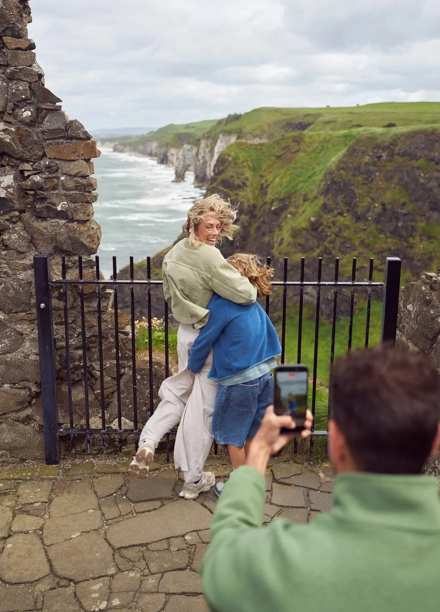 A couple embrace for a photograph at a coastal viewpoint overlooking rugged green cliffs and the sea, while a man in the foreground captures the moment on his phone.