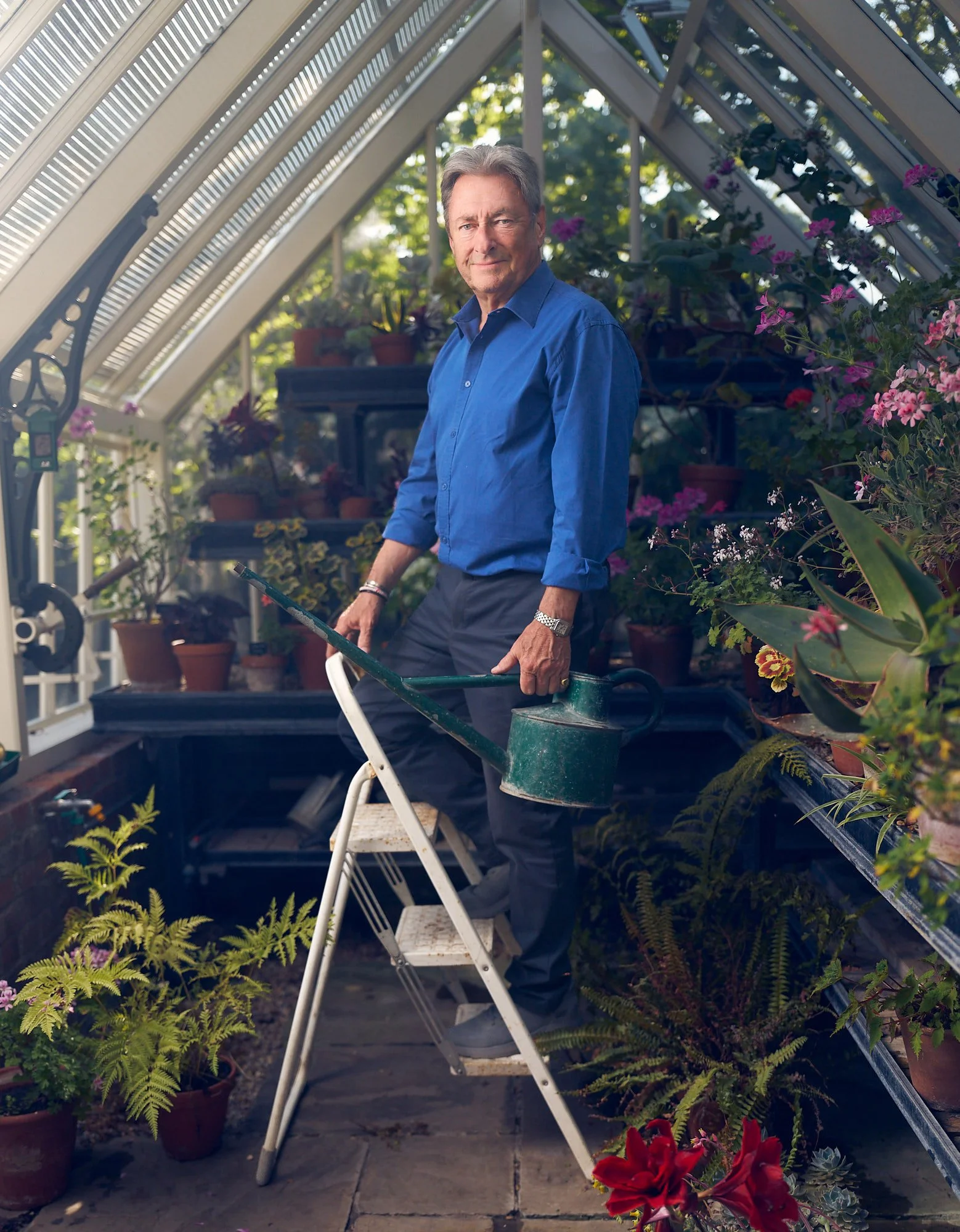 Alan Titchmarsh standing in a greenhouse surrounded by flowering plants, holding a green watering can beside a set of potting steps.