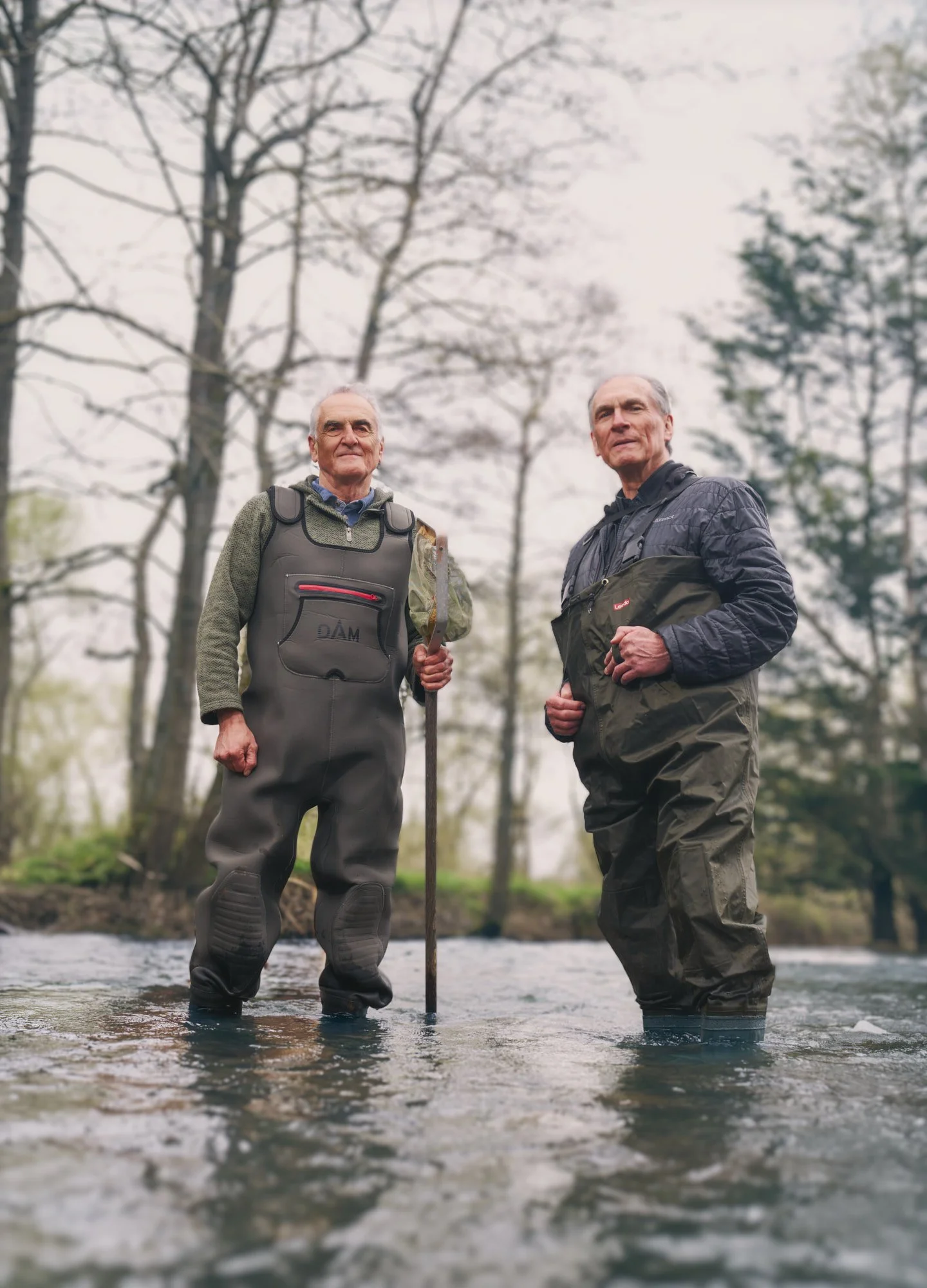 Two men wearing chest waders stand in a shallow river surrounded by woodland, one holding a long wooden pole, during an outdoor conservation project.