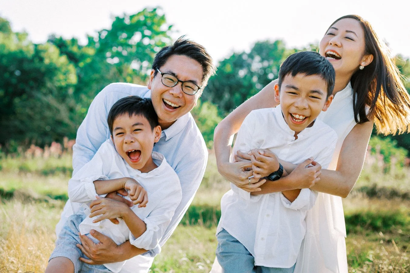 Family of four laughing and playing outdoors in a sunny park, with two children and two adults, all smiling during a family photoshoot in Hampstead Heath by the best North London family photographer Otalia Onta.