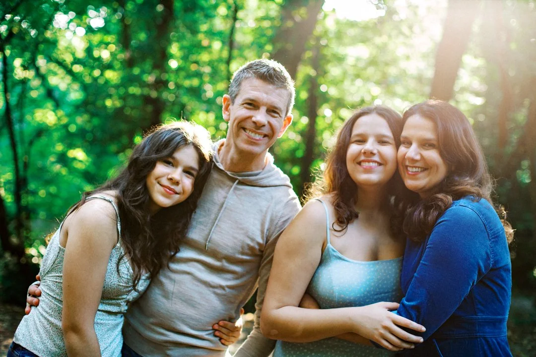 A happy family of four smiling and posing outdoors in a forest with sunlight filtering through the trees in Hampstead Heath during their North London family photography session