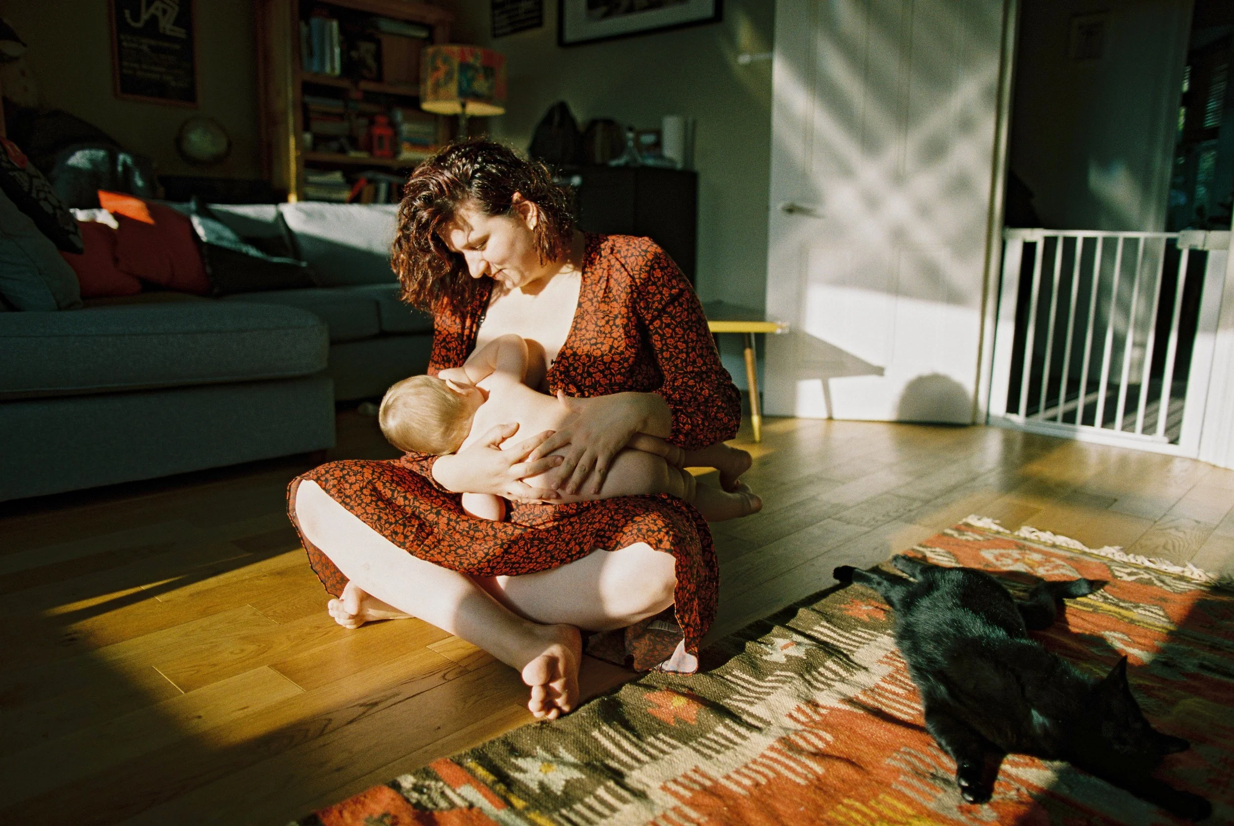A mother breastfeeding her baby bathed in winter light whilst the cat lounges nearby, portrait taken during a family photoshoot in Hampstead, London