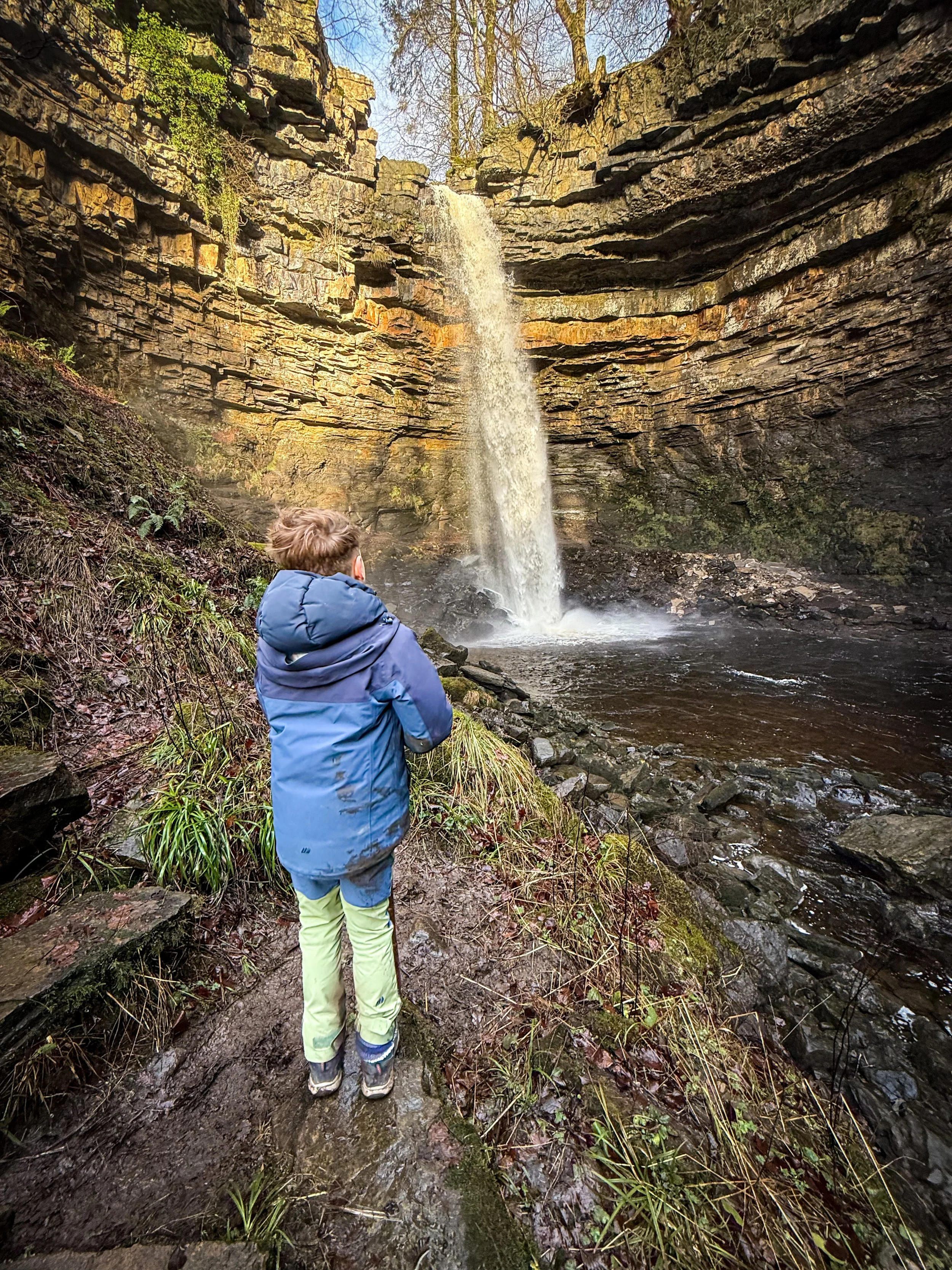 Child standing next to Hardraw force waterfall in a rocky gorge in winter