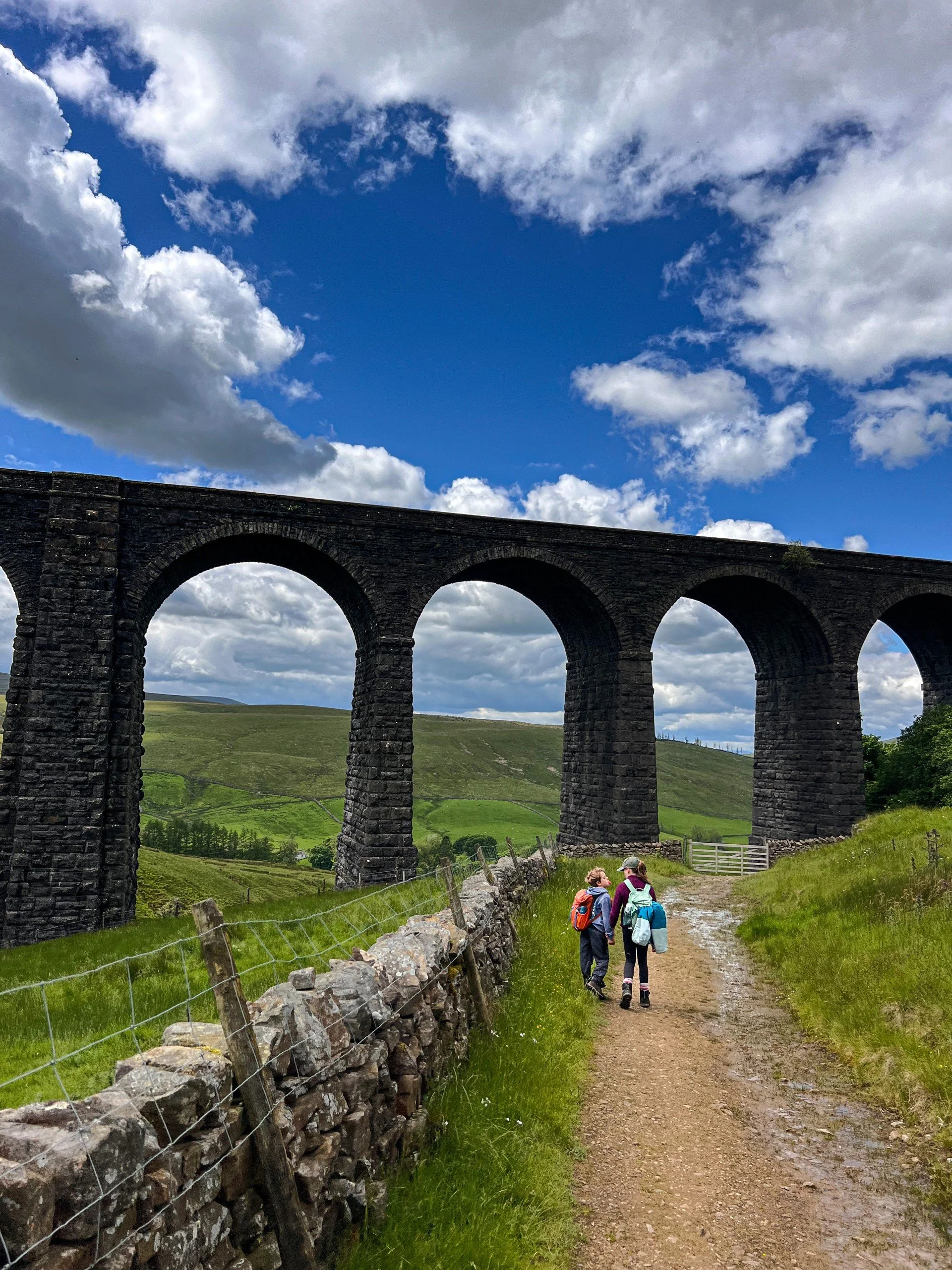 Two children walking torwards a gate under a large viaduct with blue skies ahead